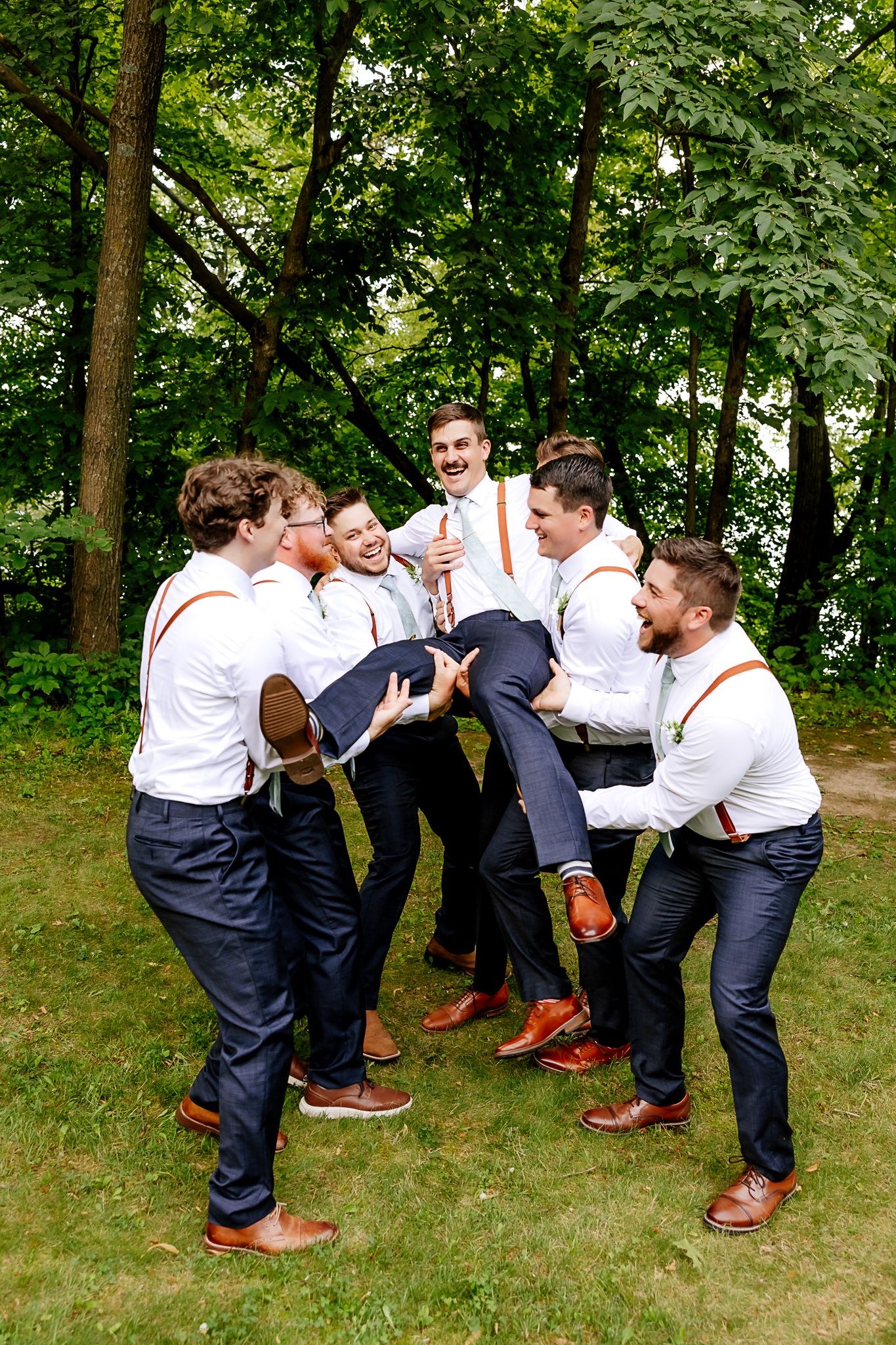 Groomsmen in white shirts and suspenders joyfully lift the groom outdoors at a wedding.