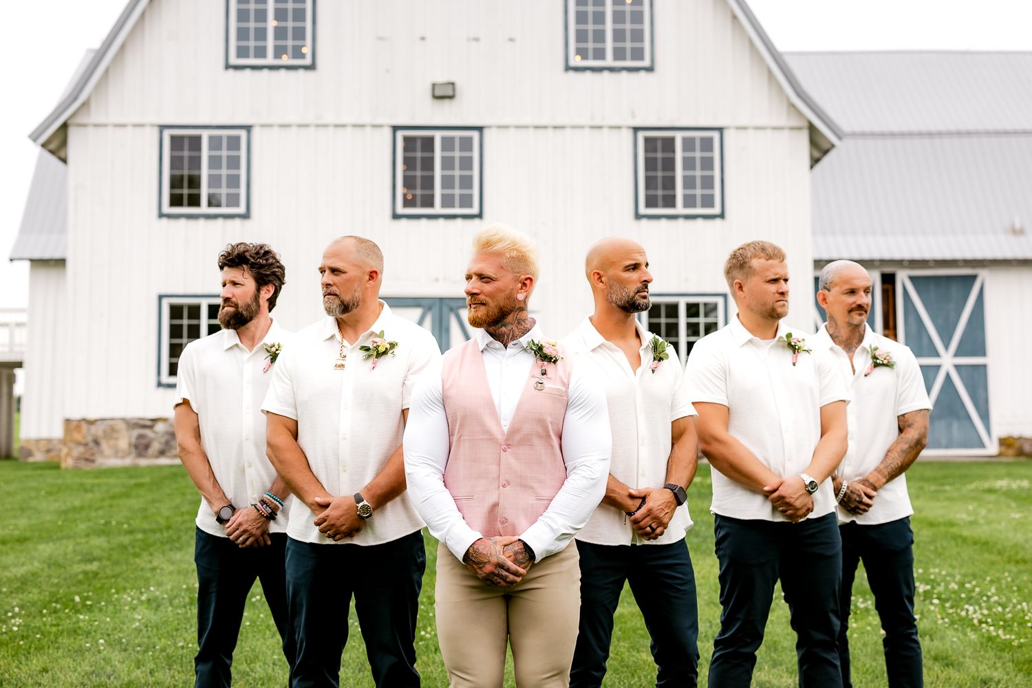 Groom in pink vest and white shirt stands with five groomsmen before a white barn on a sunny wedding day.