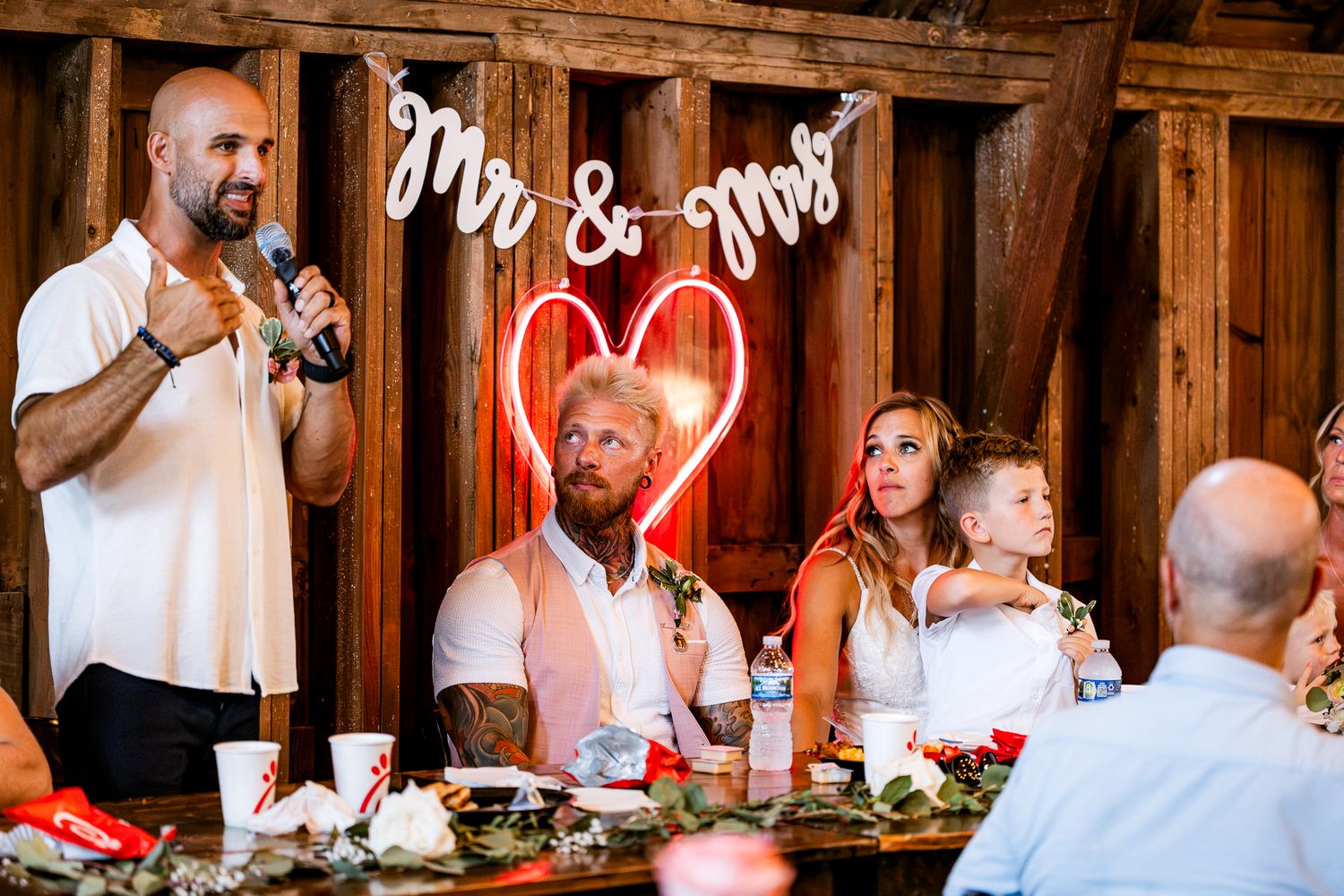 Man giving speech at rustic wedding reception with Mr & Mrs sign and neon heart behind seated bride and groom.