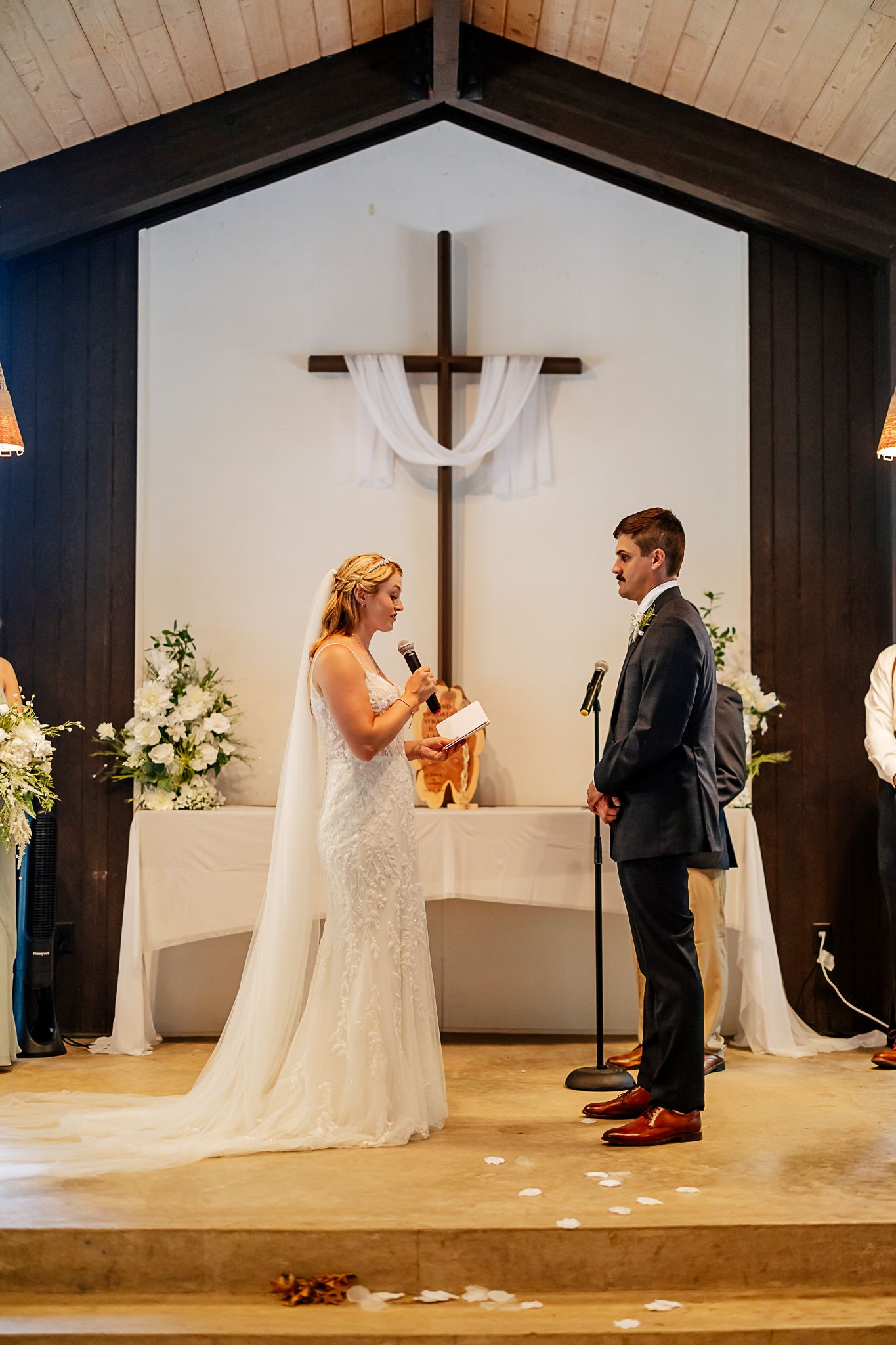 Bride reading vows to groom at church altar with wooden cross and white floral arrangements.