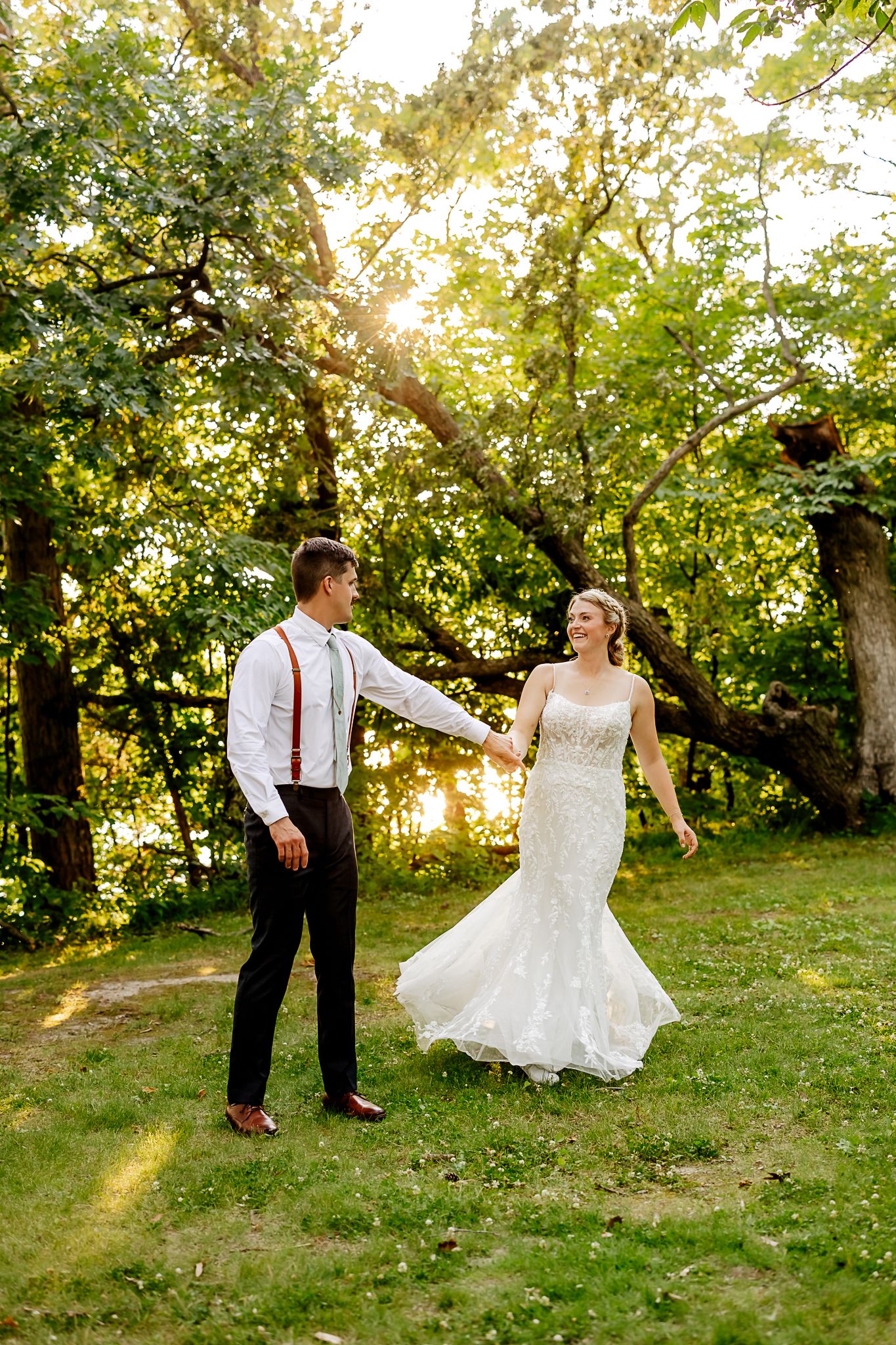 Bride and groom hold hands dancing outdoors in golden sunlight among lush green trees in wedding attire.