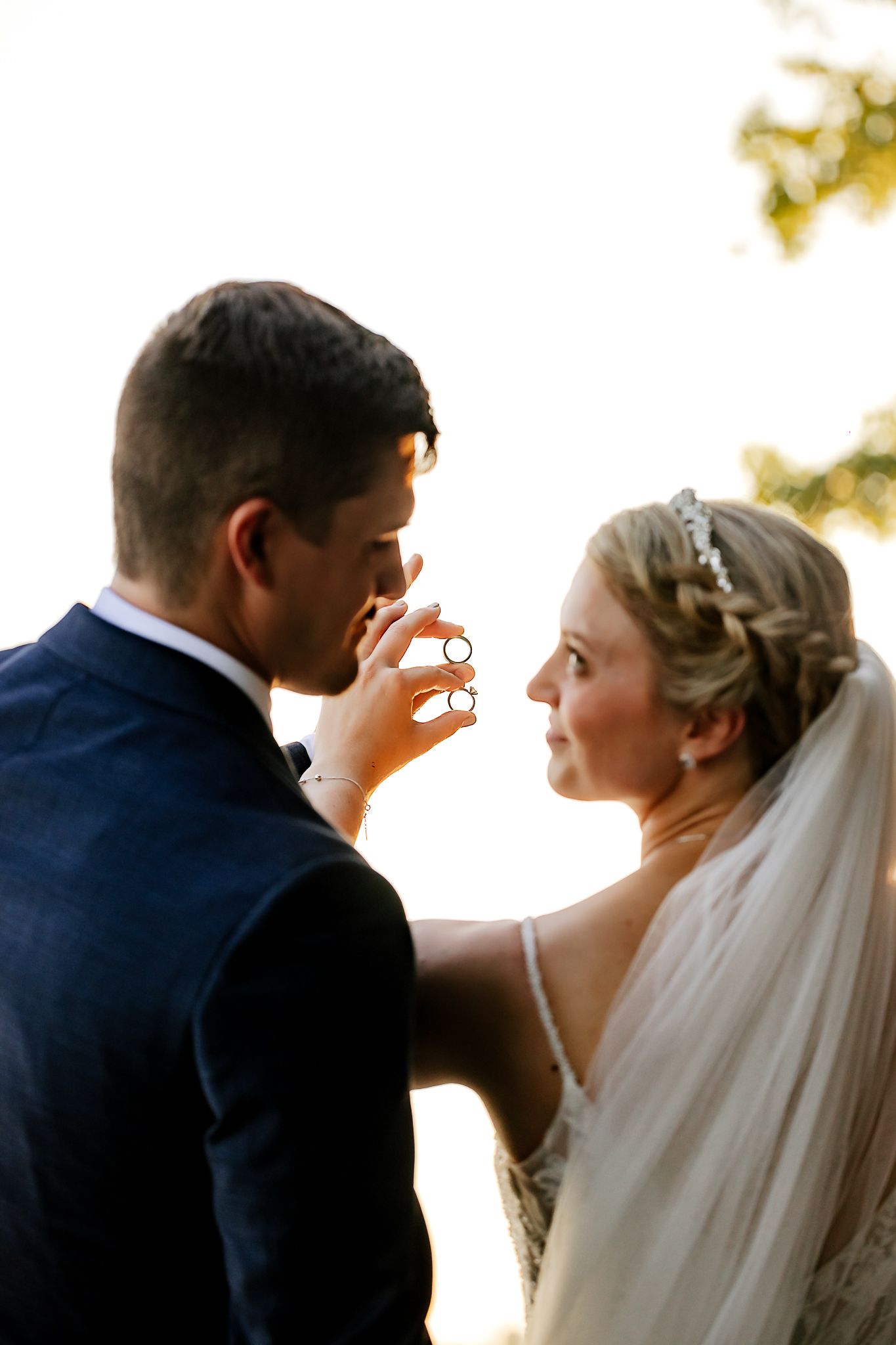 Bride and groom holding wedding rings together in a romantic backlit outdoor portrait on their wedding day.