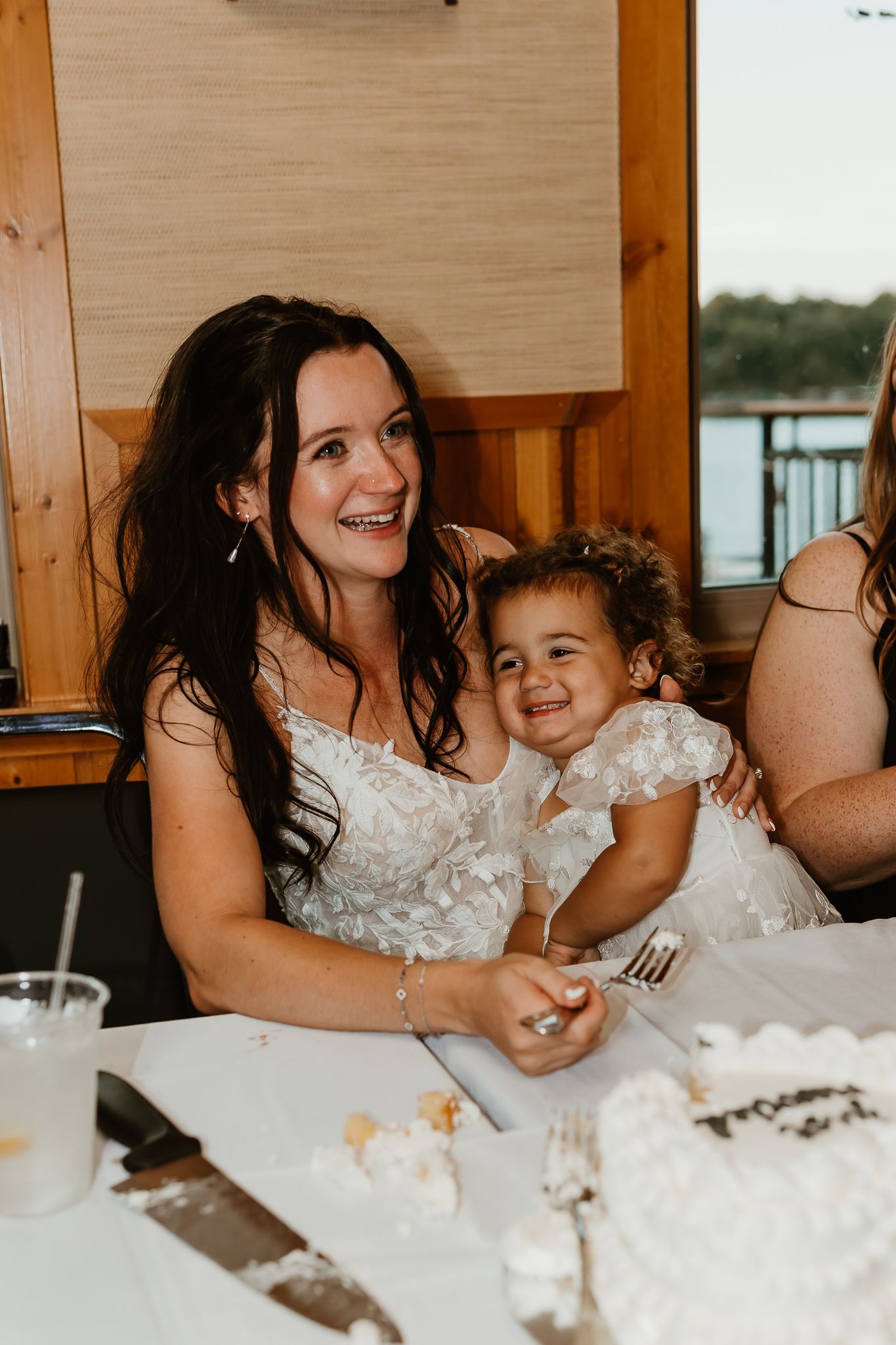 Smiling bride in a white lace dress holds a laughing toddler on her lap beside a wedding cake and cake knife at a lakeside reception.