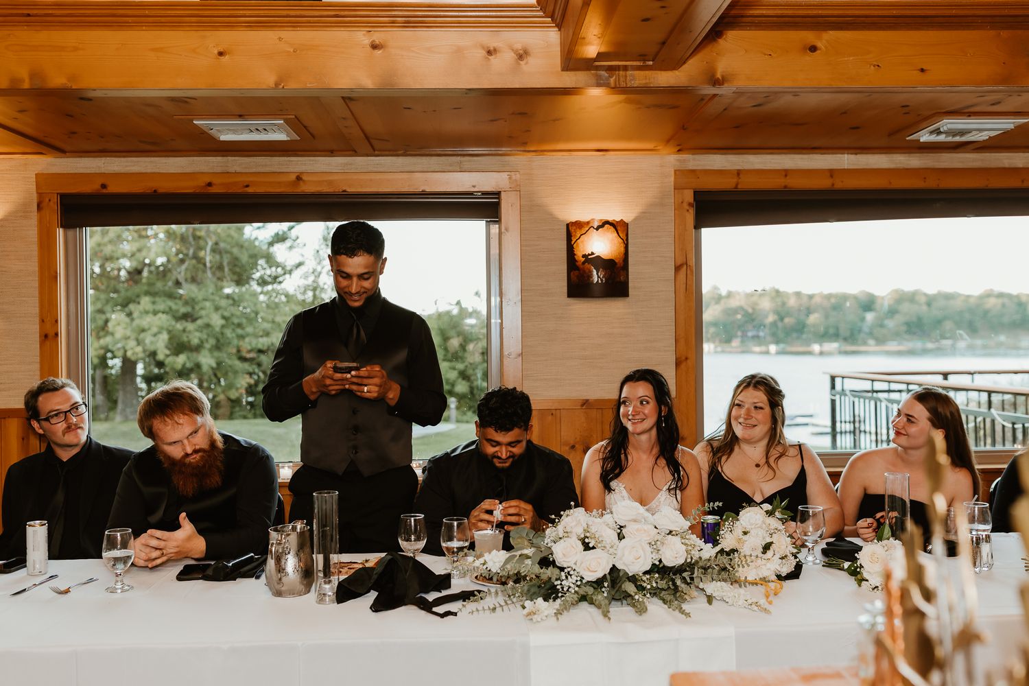 Wedding guests seated at a long table with white floral centerpieces while a man stands to give a toast in a lakeside venue.