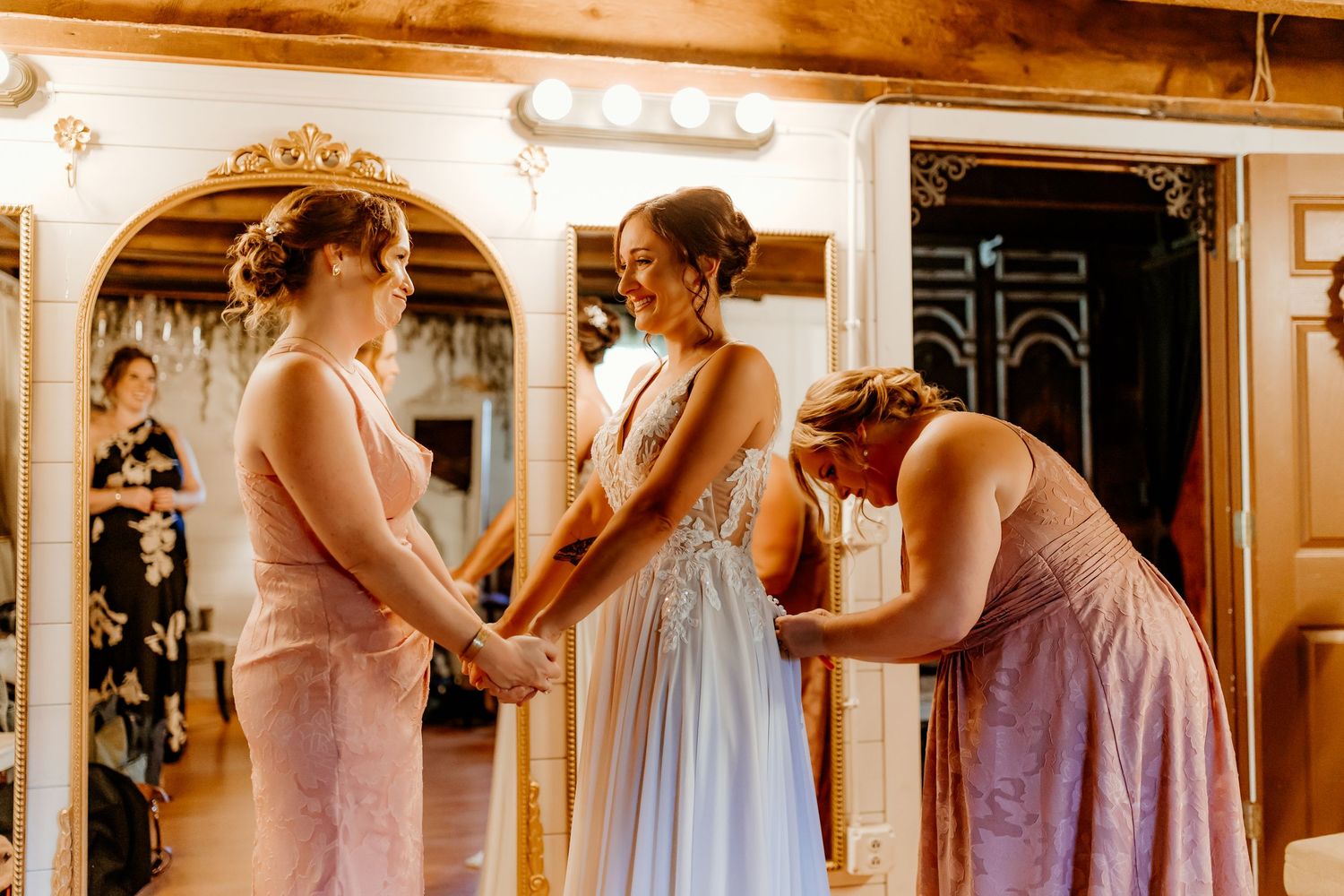 Bride in a white gown holds hands with two bridesmaids in blush pink dresses under warm lights in a rustic wedding venue.