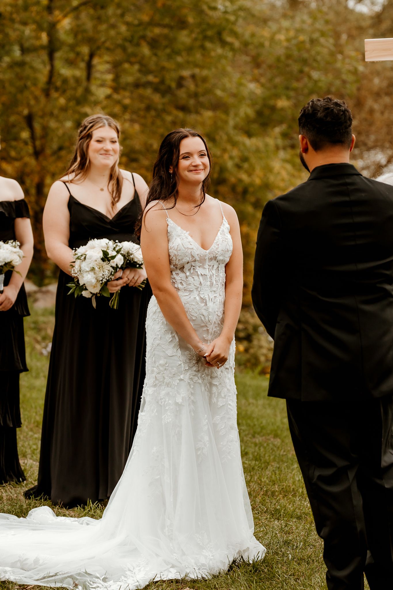 Smiling bride in a lace wedding gown holds her hands together during an outdoor ceremony as a bridesmaid in black holds a white bouquet.