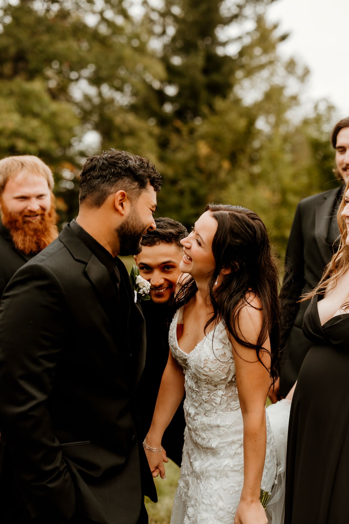 Smiling bride in a lace wedding dress laughs with groomsmen in black suits during an outdoor wedding celebration among evergreen trees.