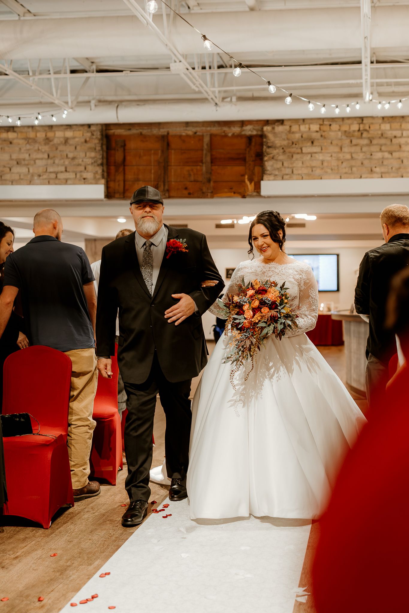 Bride in a white gown with an autumn bouquet walks down the aisle on a white runner, arm in arm with a man in a dark suit and cap.