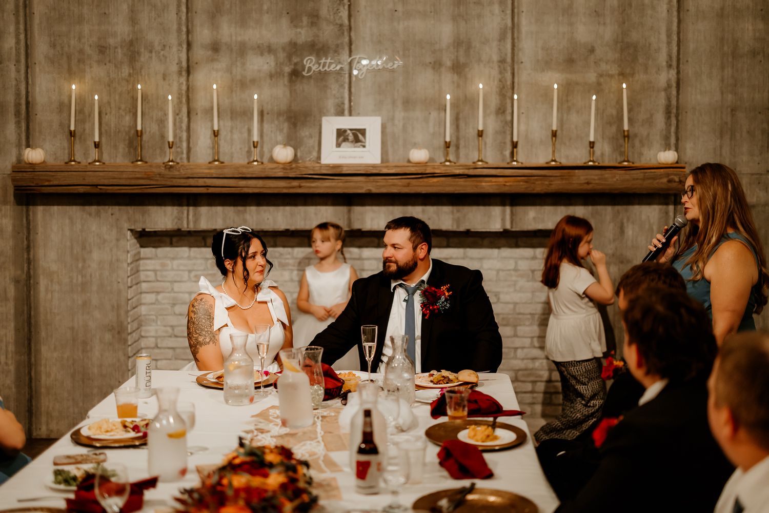 A bride and groom smile at their head table with a young girl as a woman gives a toast at a rustic candlelit wedding reception.