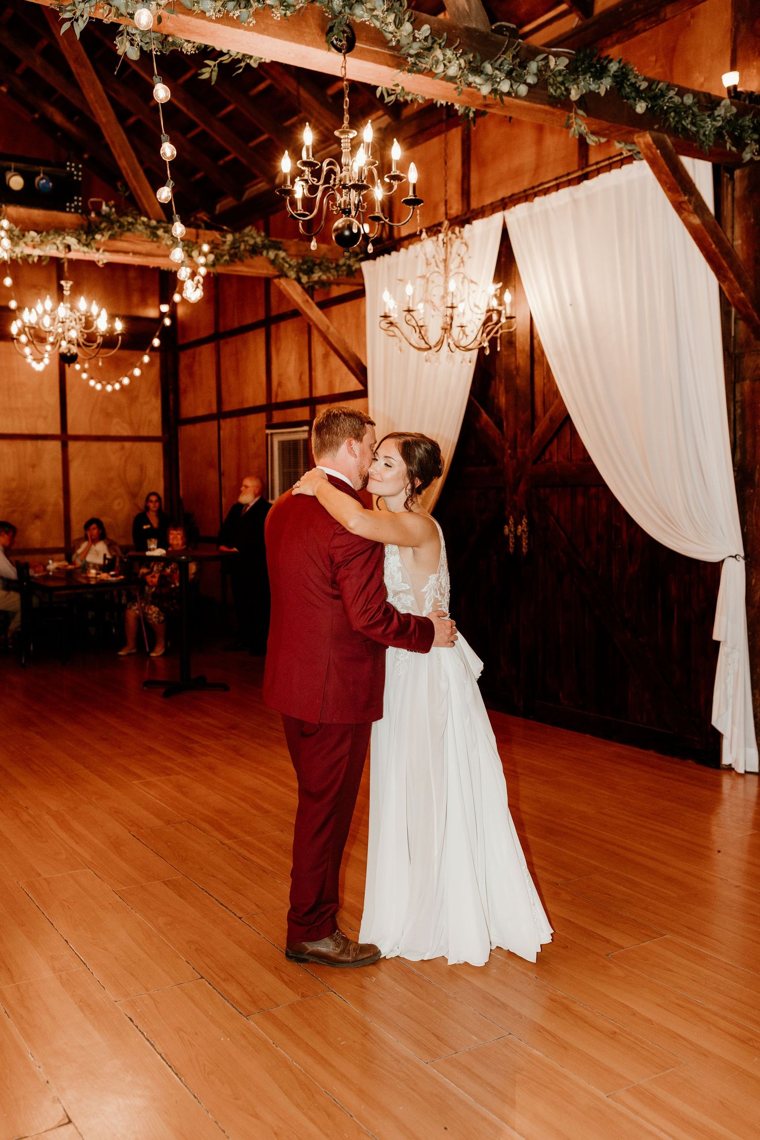 Bride in a white gown and groom in a burgundy suit share a first dance in a rustic barn wedding venue with chandeliers and draped curtains.