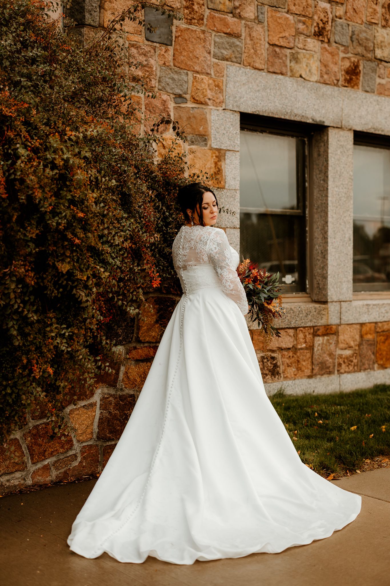 Bride in a white lace-sleeved A-line wedding gown holds a bouquet beside a stone building and autumn foliage.