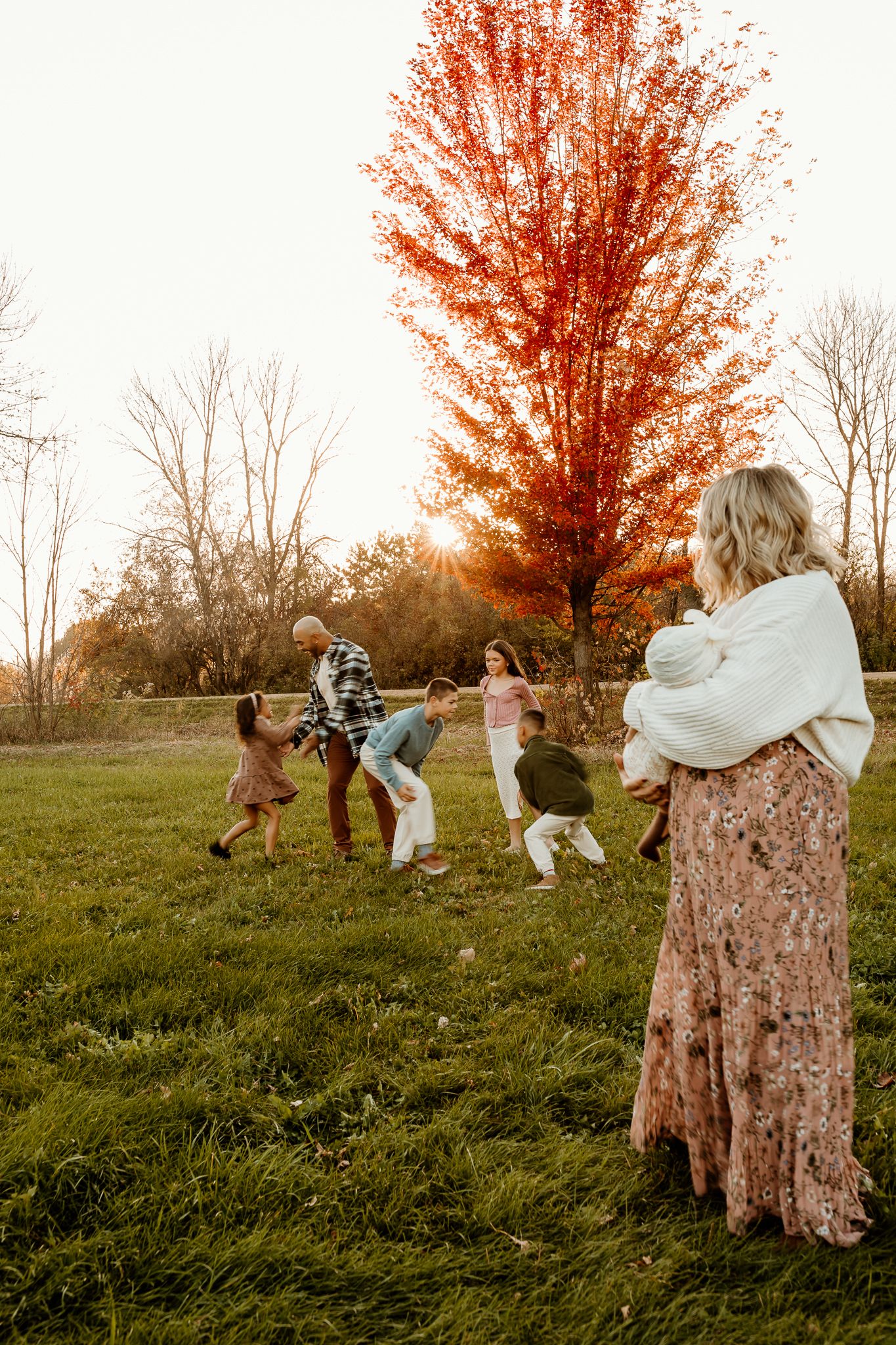 Family playing together on a grassy field at sunset beside a vibrant red autumn tree while a woman holding a baby watches.