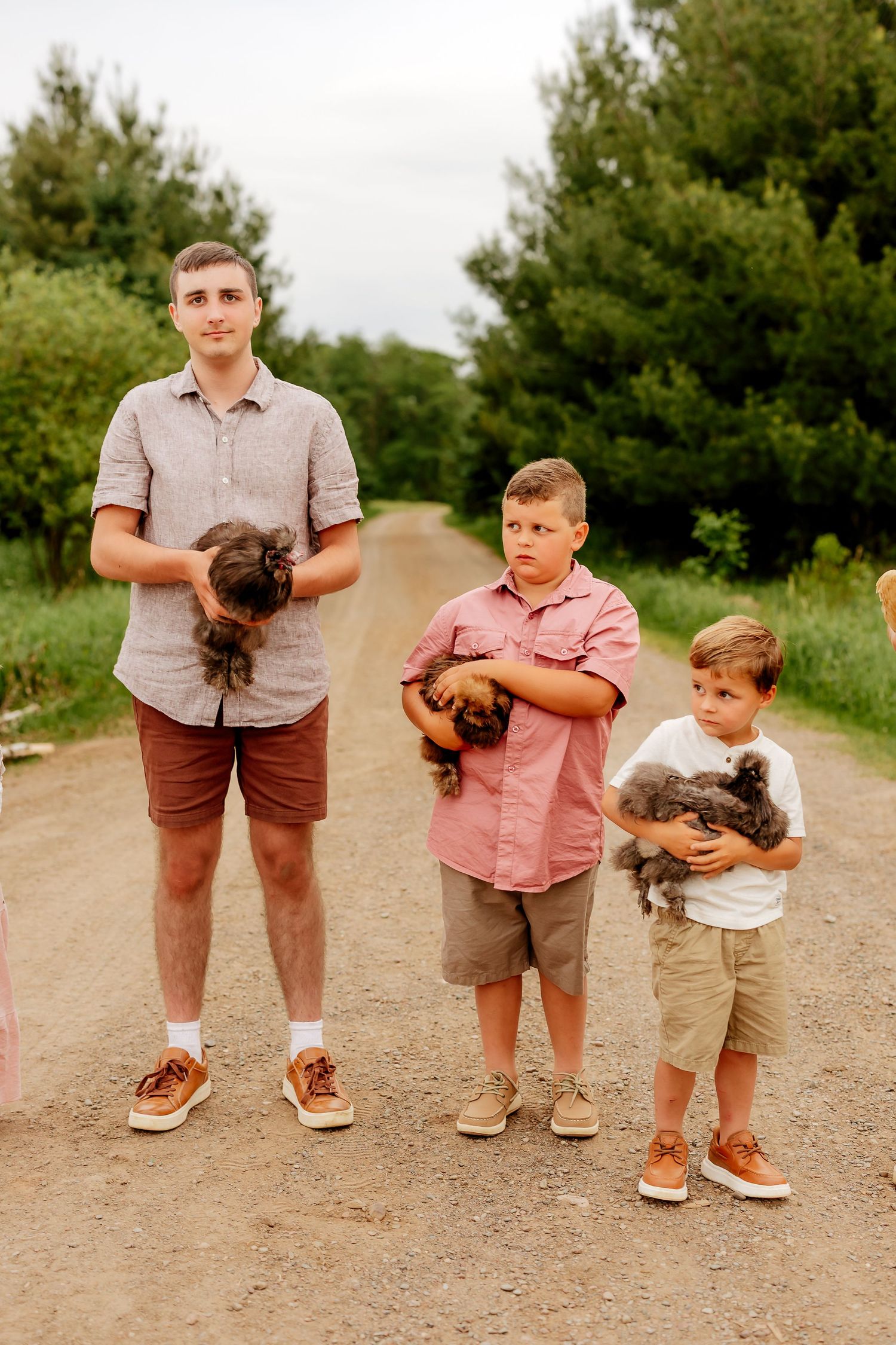 Three boys stand on a dirt road holding small animals, dressed in coordinating casual outfits outdoors.