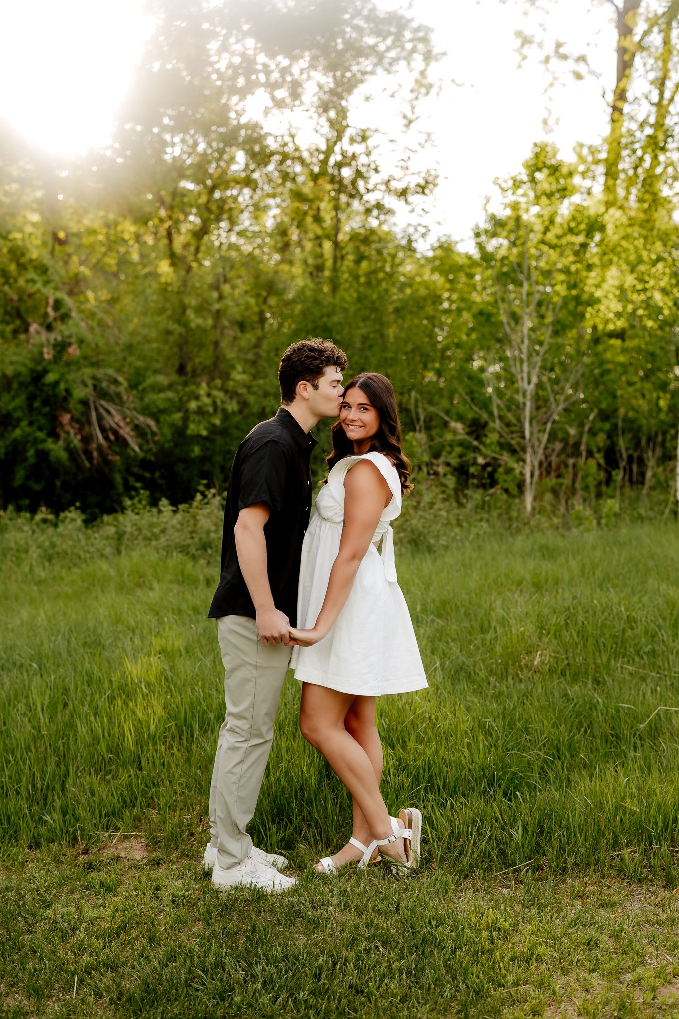 Couple embraces in lush green meadow at golden hour, man kissing woman on cheek.