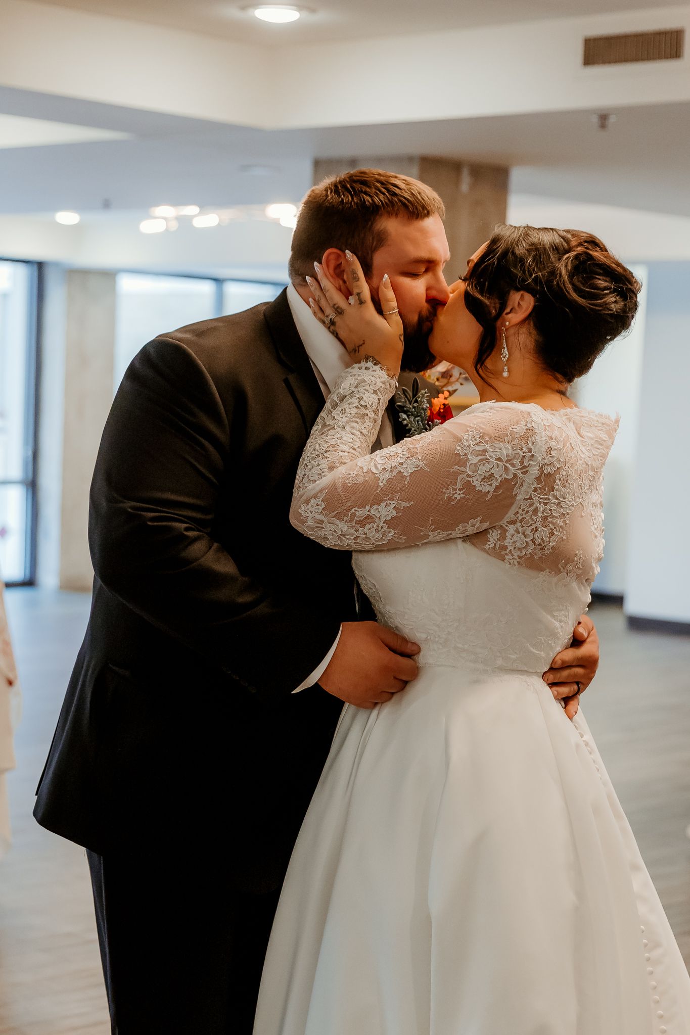Bride in a lace-sleeved white wedding gown and groom in a dark suit share an emotional kiss during their wedding ceremony indoors.