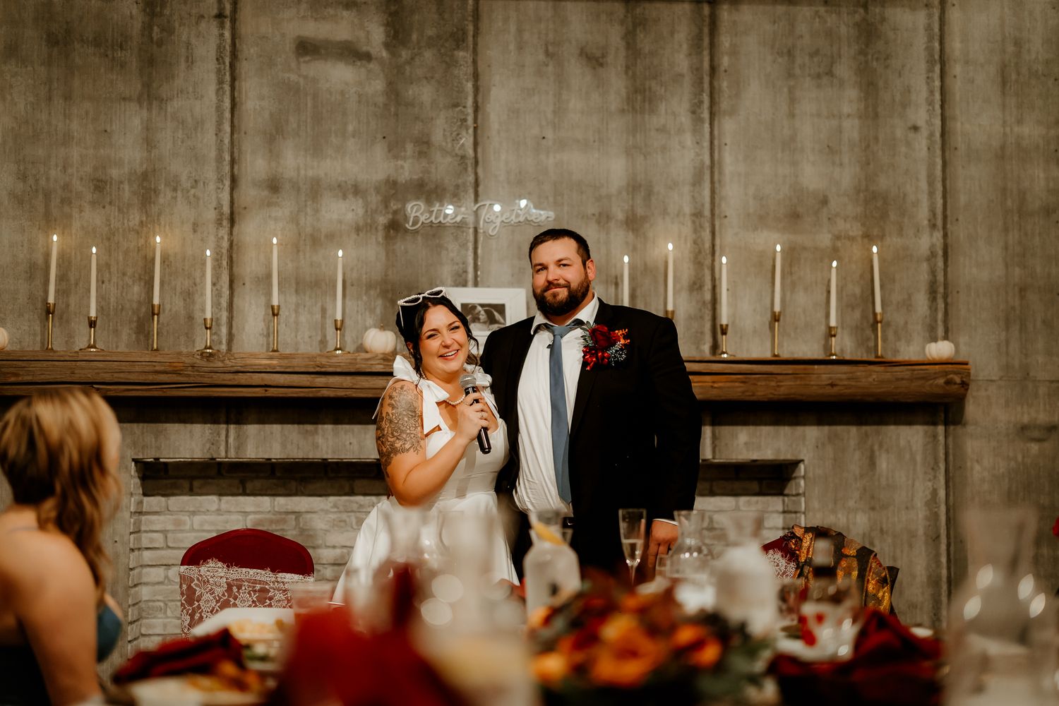 Smiling bride in a white dress speaks into a microphone beside her groom at a rustic wedding reception with candles and autumn floral center