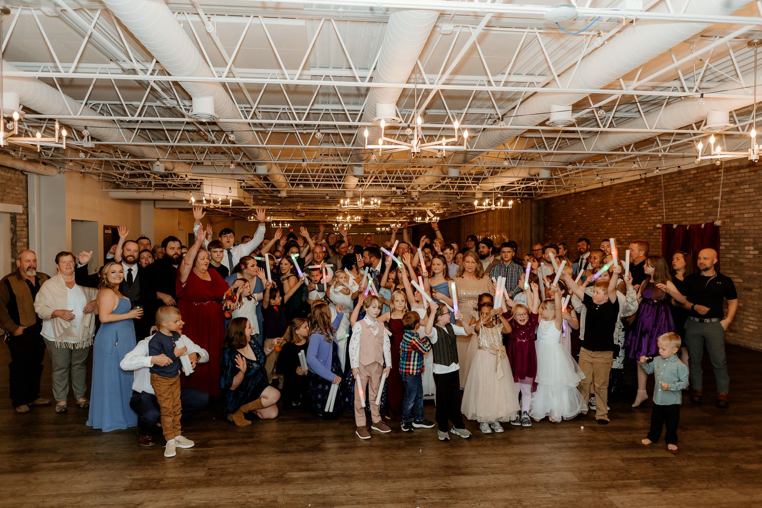 Large group of wedding guests, including children in formal dresses, cheering and posing together in a rustic venue with exposed brick and s