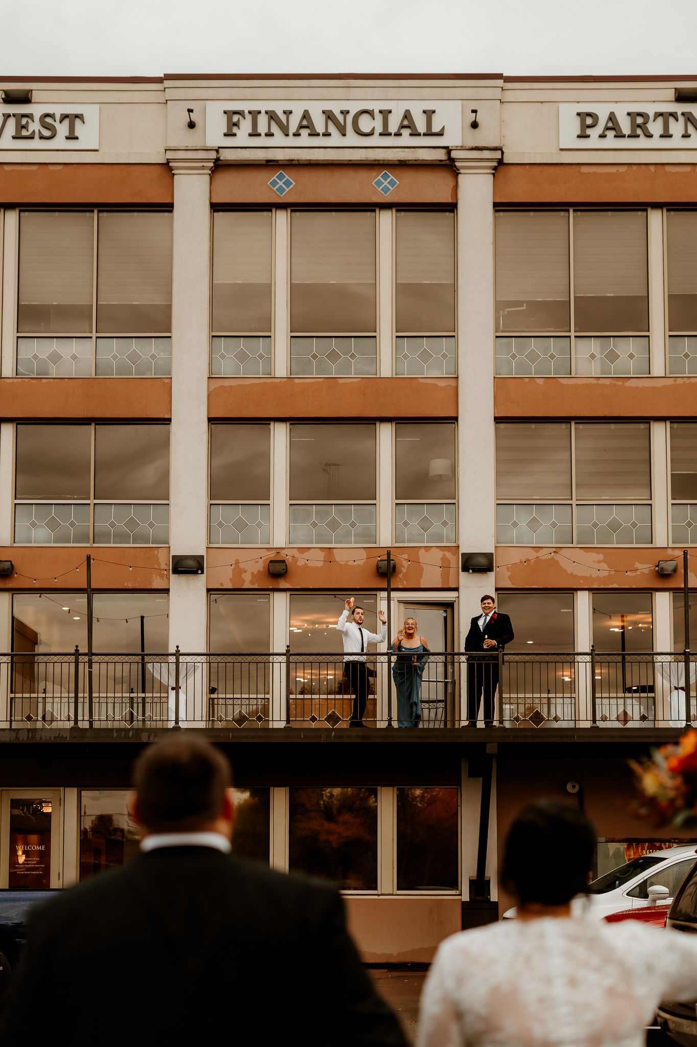 Wedding guests watch from the street as three people stand on the balcony of a brick Financial Partners building at dusk.