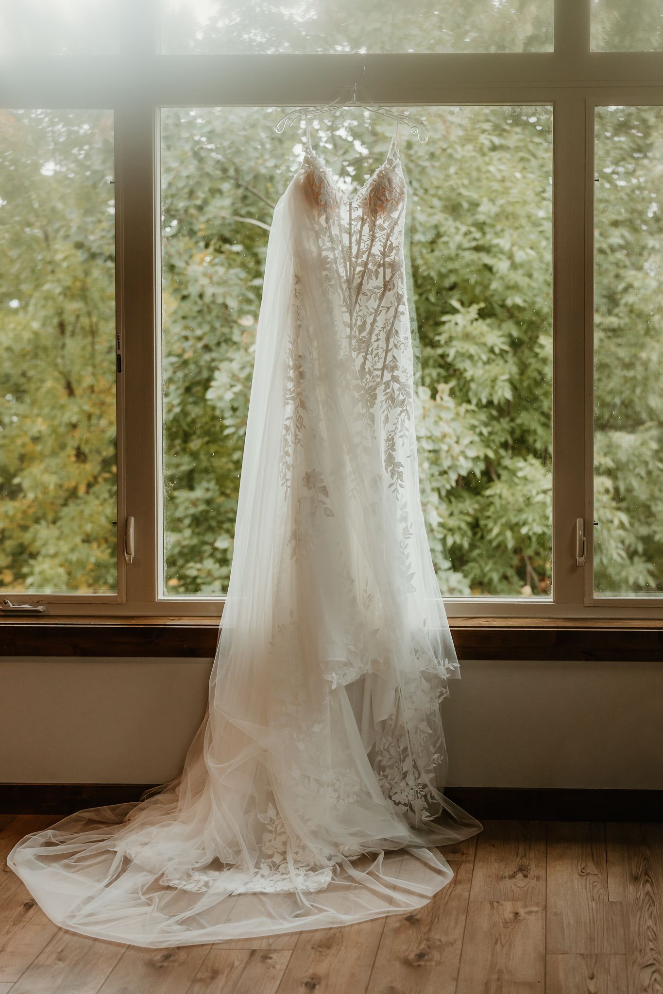 White lace wedding dress with a long sheer veil hanging by a large window overlooking green trees, with the train on a wood floor.