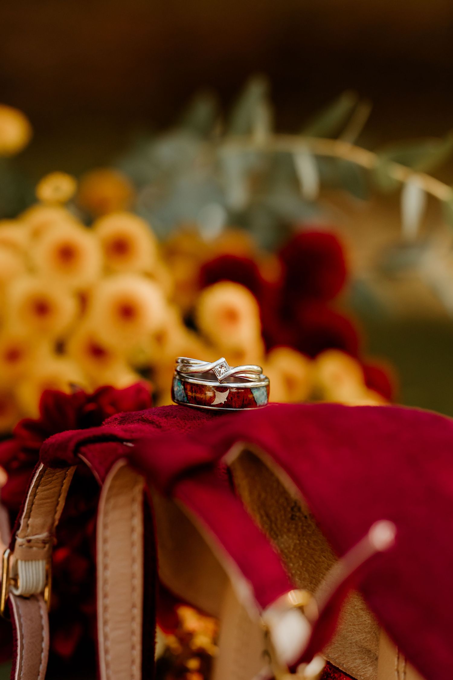 Wedding rings resting on a burgundy velvet pouch with a leather strap, surrounded by golden yellow and deep red autumn flowers.