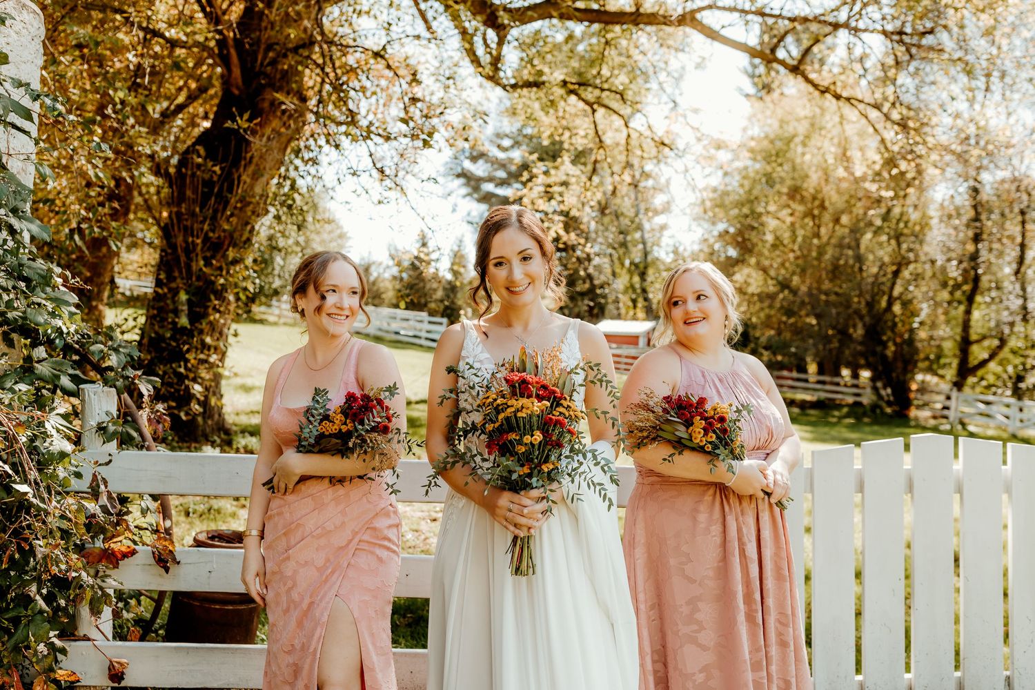 Bride in a white gown stands between two bridesmaids in blush dresses, each holding colorful autumn bouquets beside a white fence.