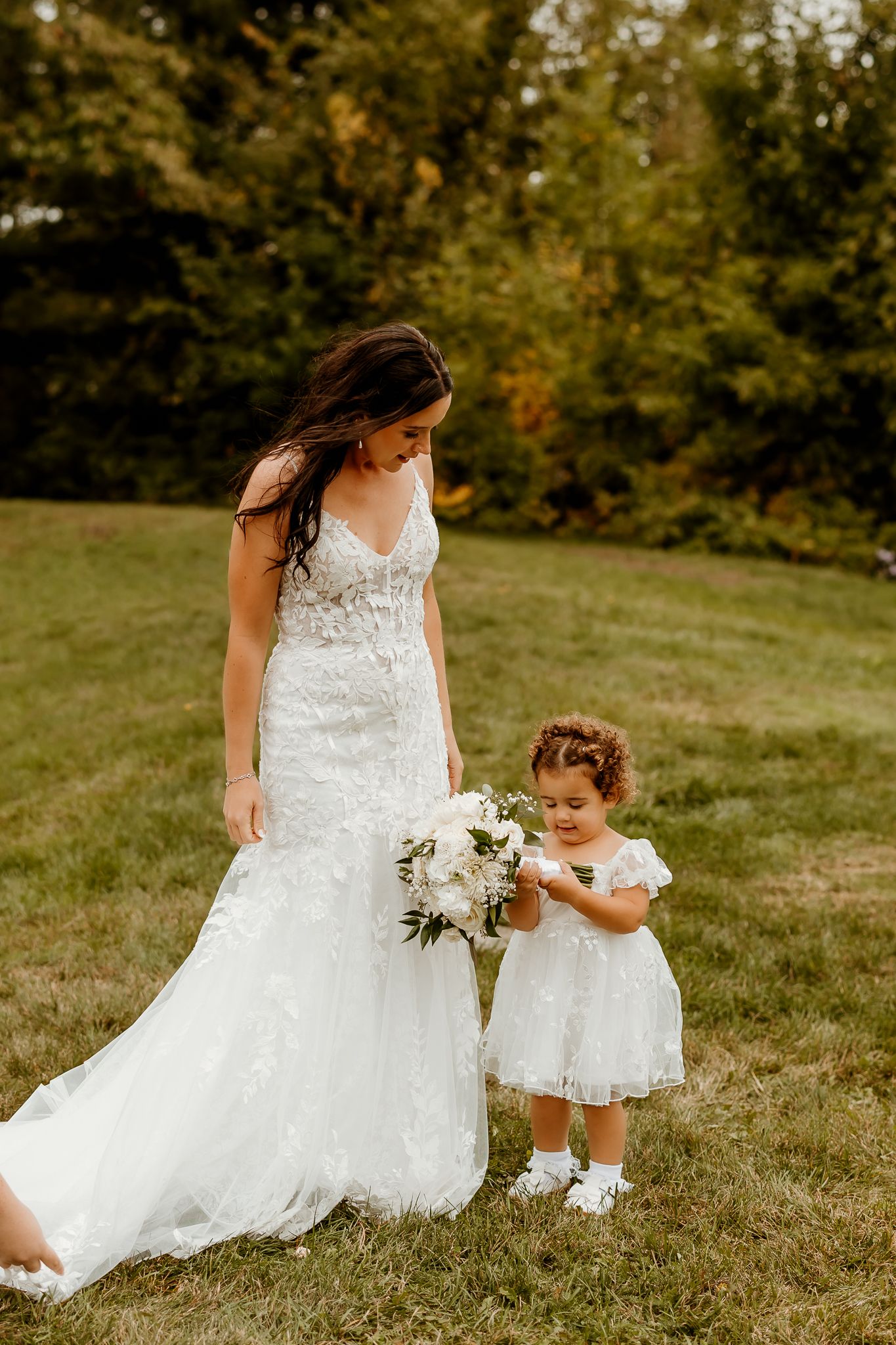 A bride in a lace wedding gown stands on a grassy lawn beside a flower girl in a white dress, who admires a white floral bouquet.
