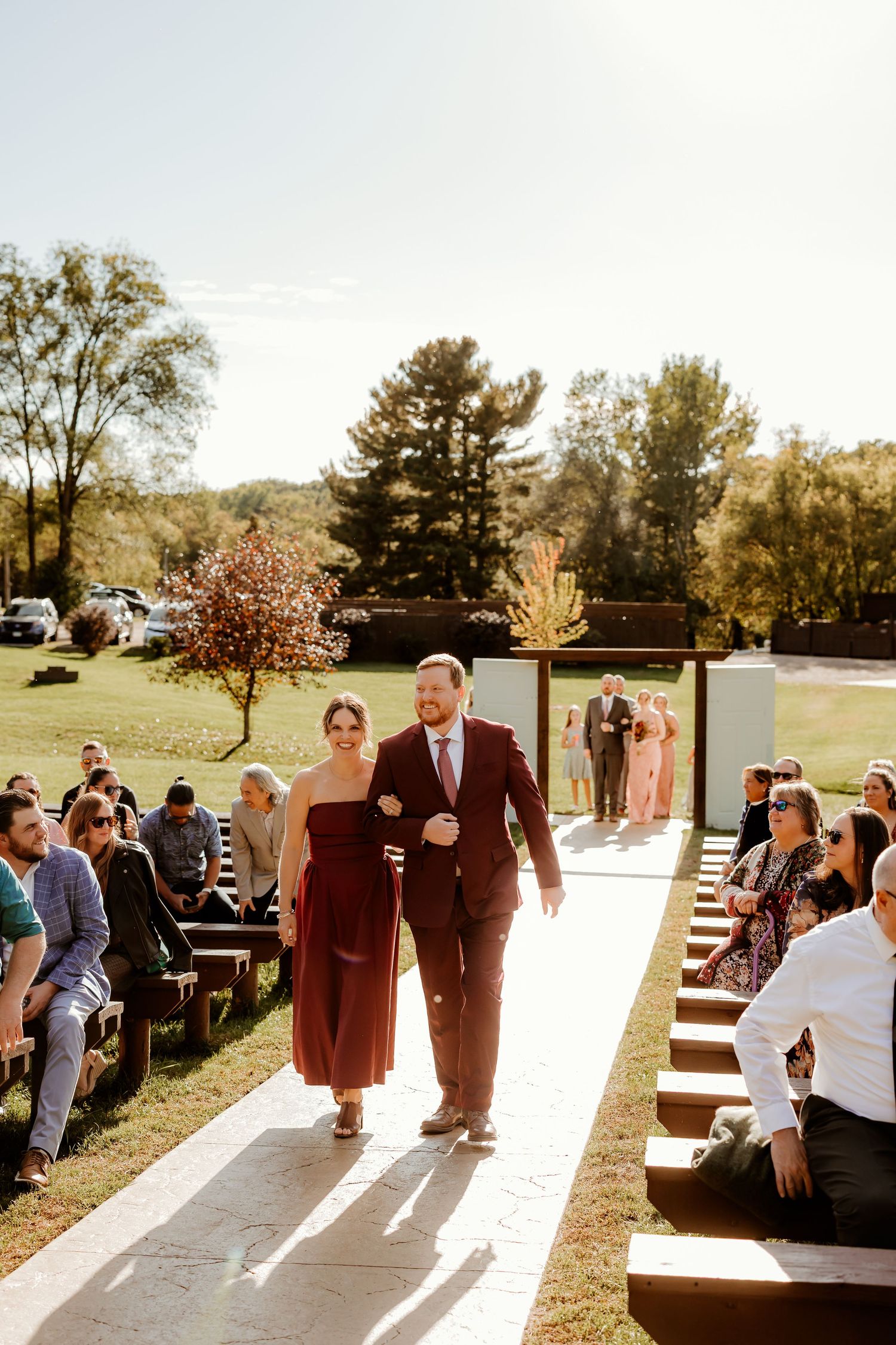 Groomsman in a burgundy suit escorts a bridesmaid in a burgundy dress down the outdoor aisle past seated guests at a sunny fall wedding.