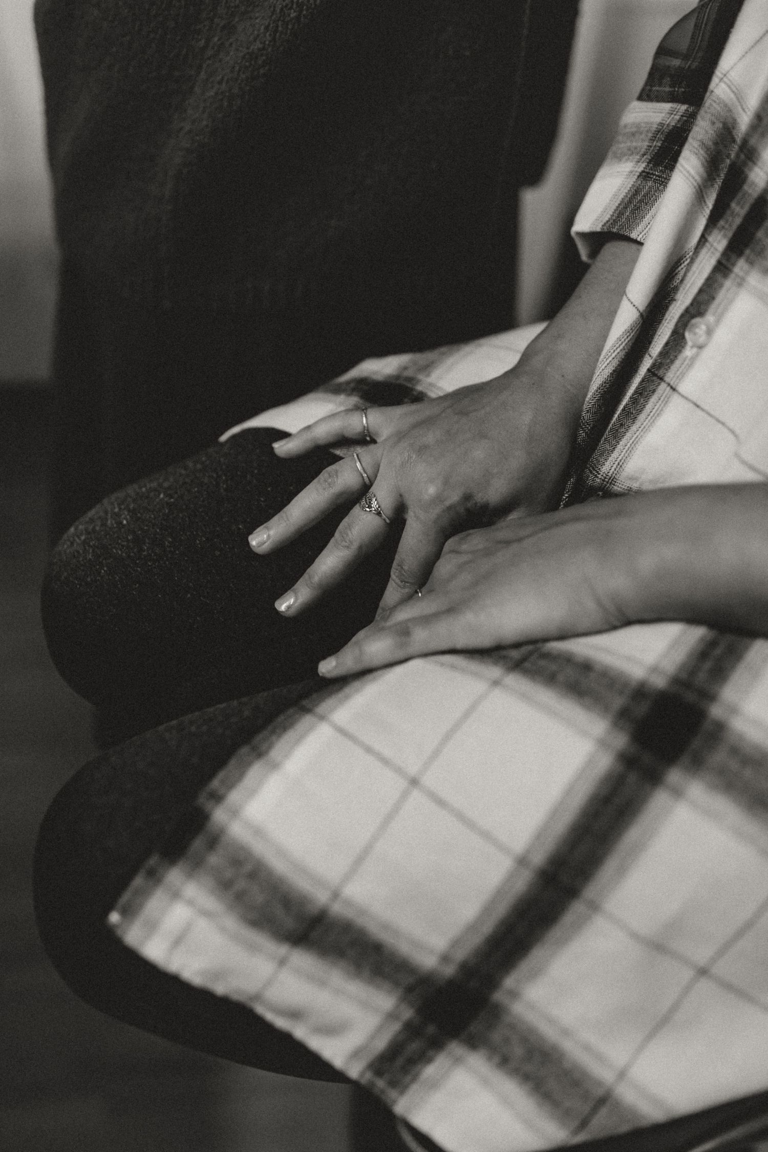 Black and white close-up of a woman's hands wearing delicate rings, resting on her knees over a plaid shirt and dark knit tights.