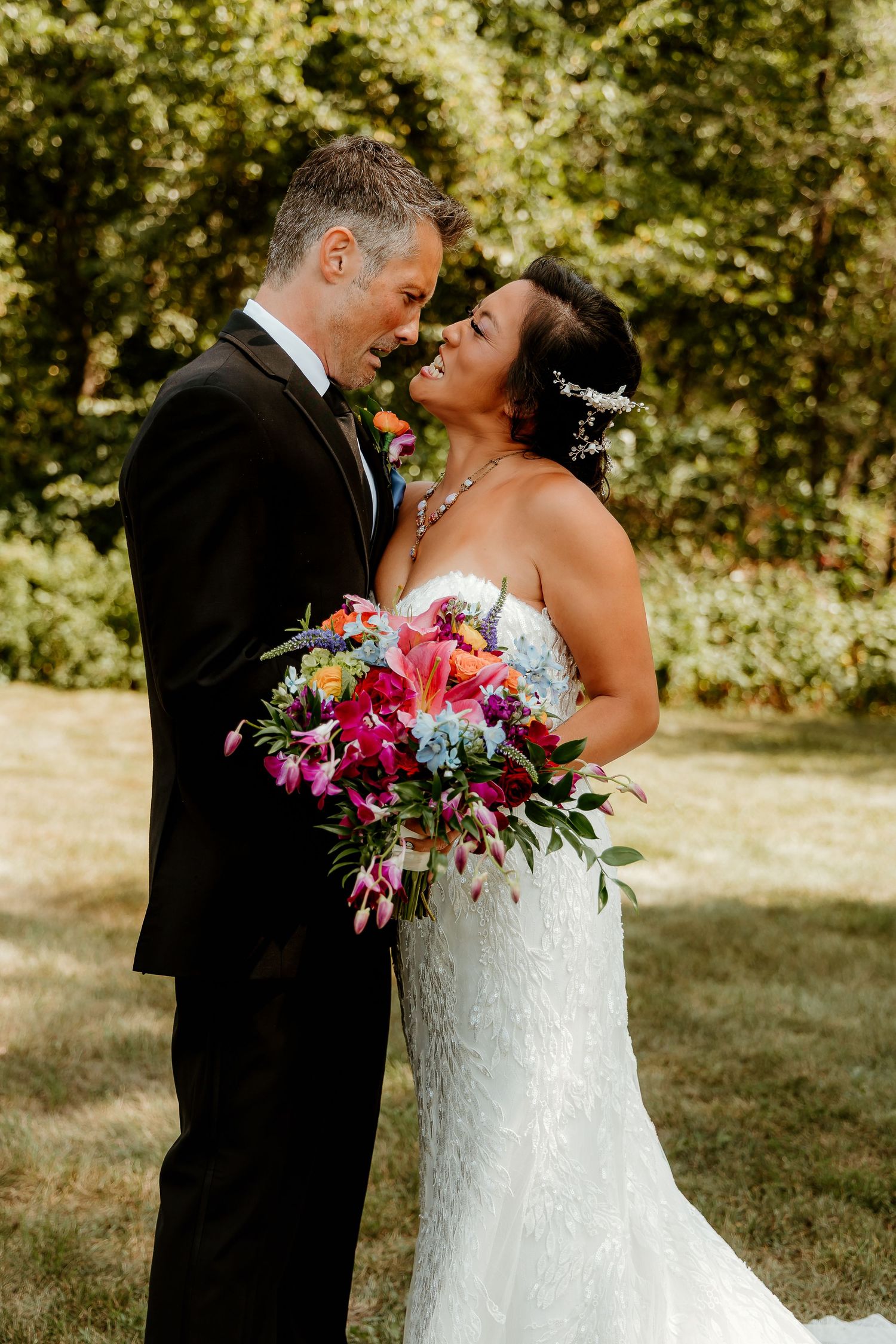 Bride and groom share a romantic moment outdoors, she holds a vibrant bouquet of pink, orange, and blue flowers.
