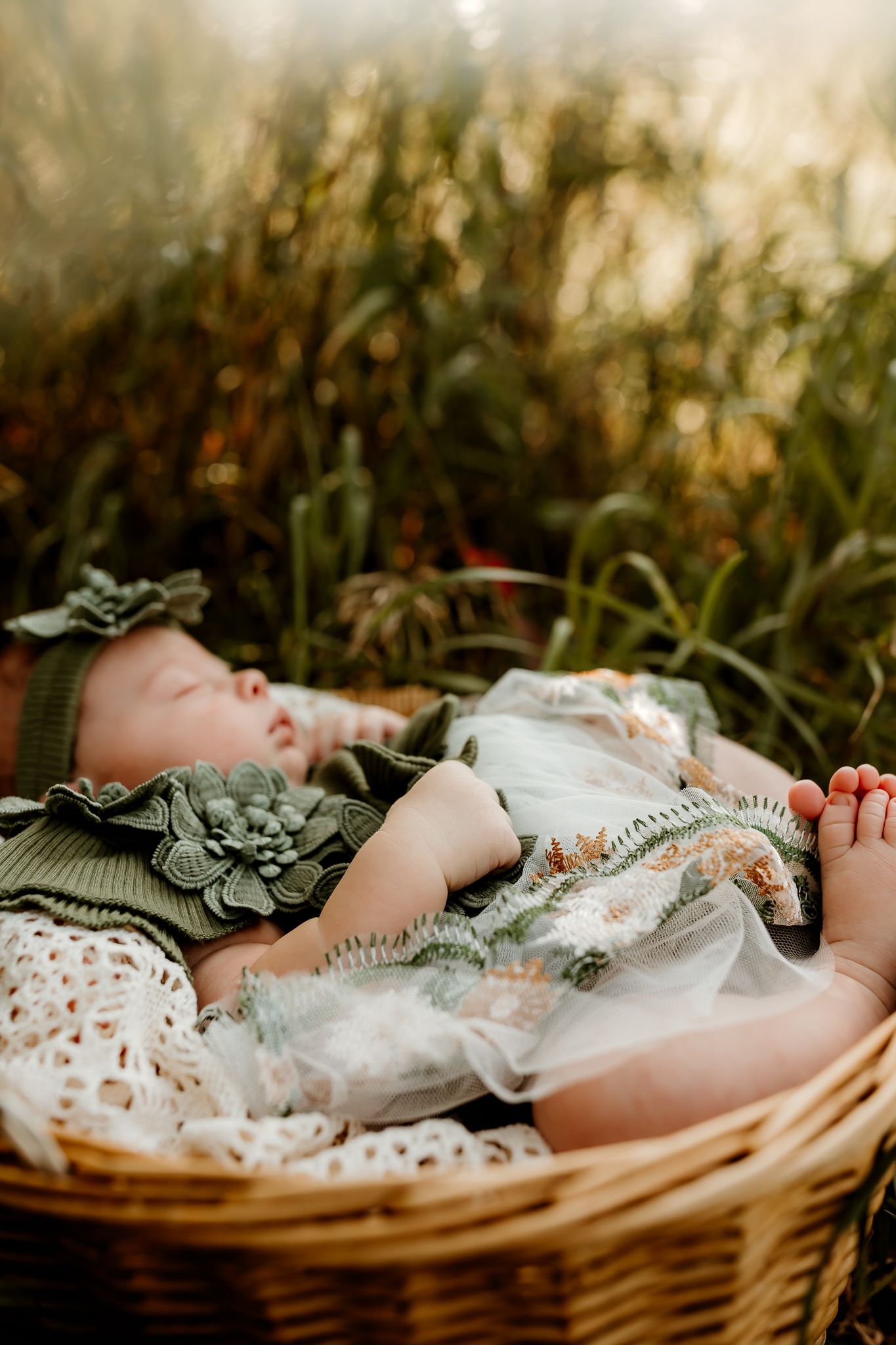 Sleeping newborn baby in green outfit and floral headband resting in a wicker basket outdoors.