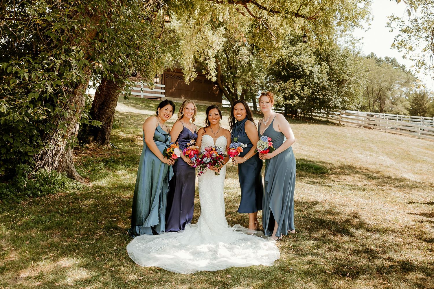 Bride in white gown with bridesmaids in dusty blue dresses holding colorful bouquets outdoors under a large tree.