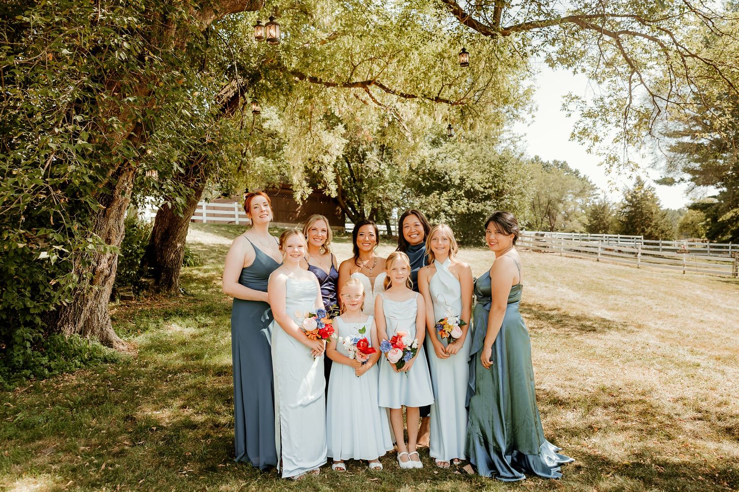 Bridal party in blue gowns holding colorful bouquets, posing under a tree on a sunny day at an outdoor wedding venue.