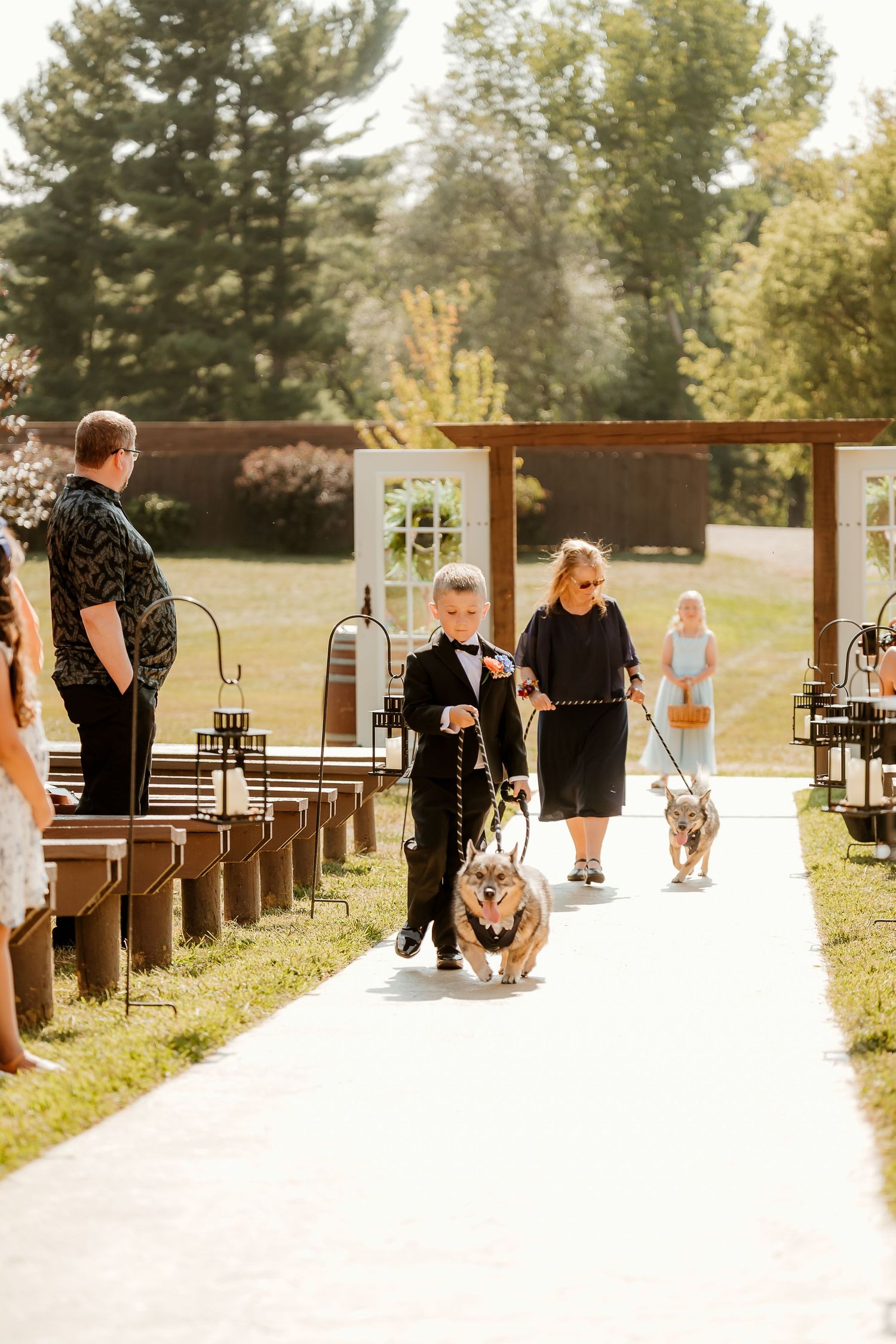 A young ring bearer in a tuxedo walks a corgi down the aisle at an outdoor wedding ceremony.