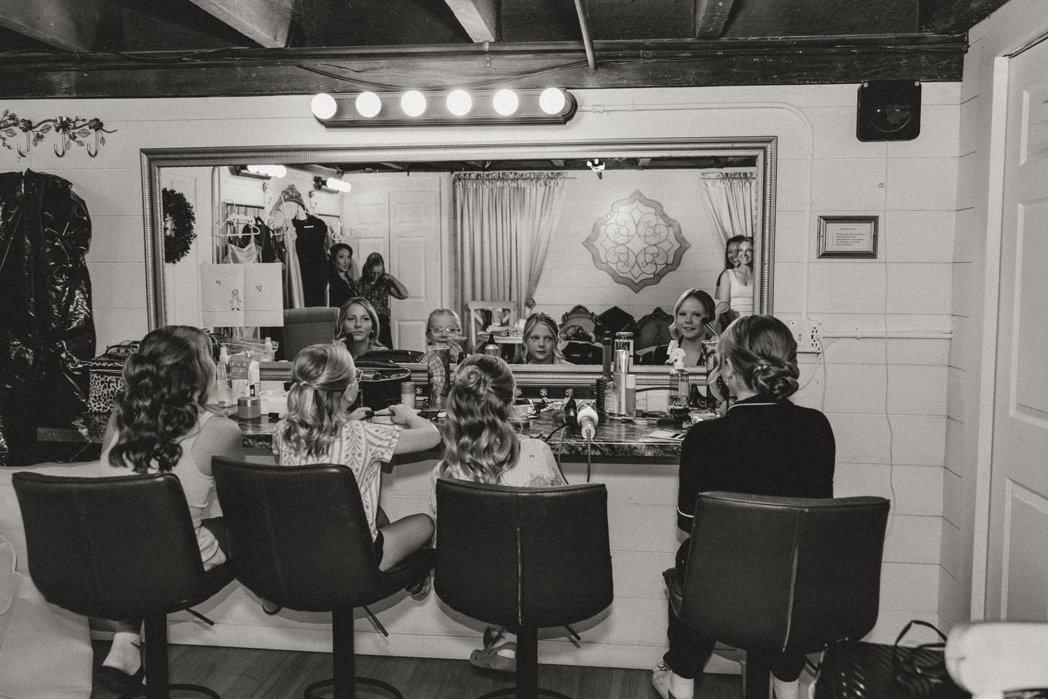 Black and white photo of women sitting at a salon mirror with bright vanity lights during a bridal party preparation.