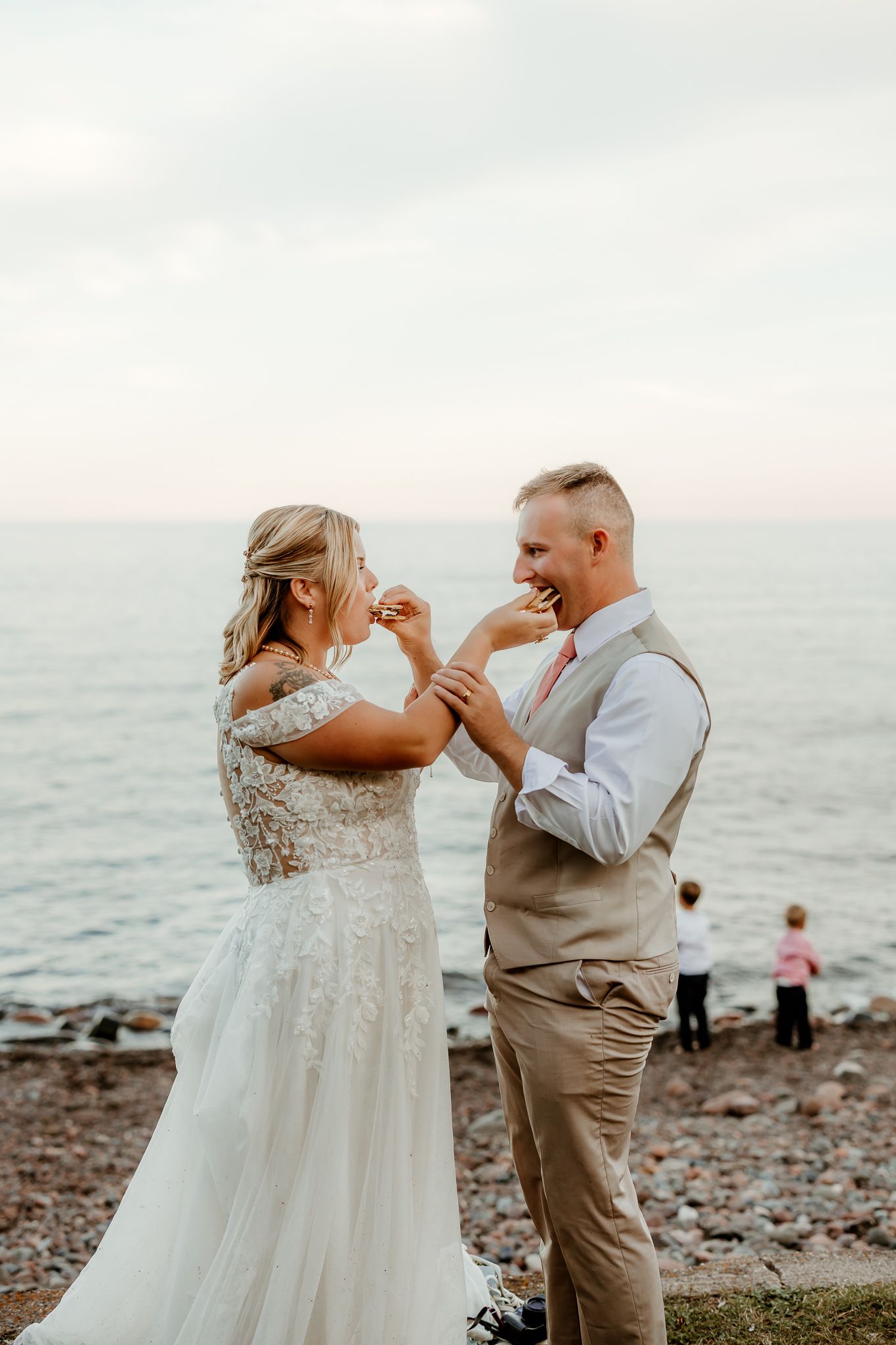 Bride and groom feeding each other cake on a rocky beach shoreline during their outdoor wedding celebration.