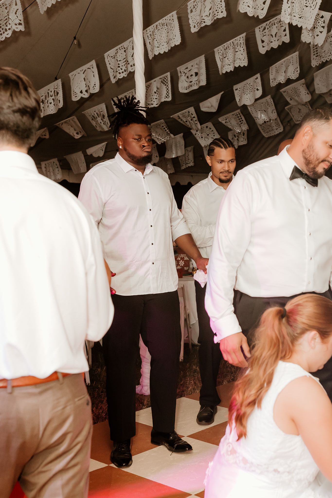 Men in white shirts and black pants socialize on a dance floor under decorative paper flags at a festive wedding reception.