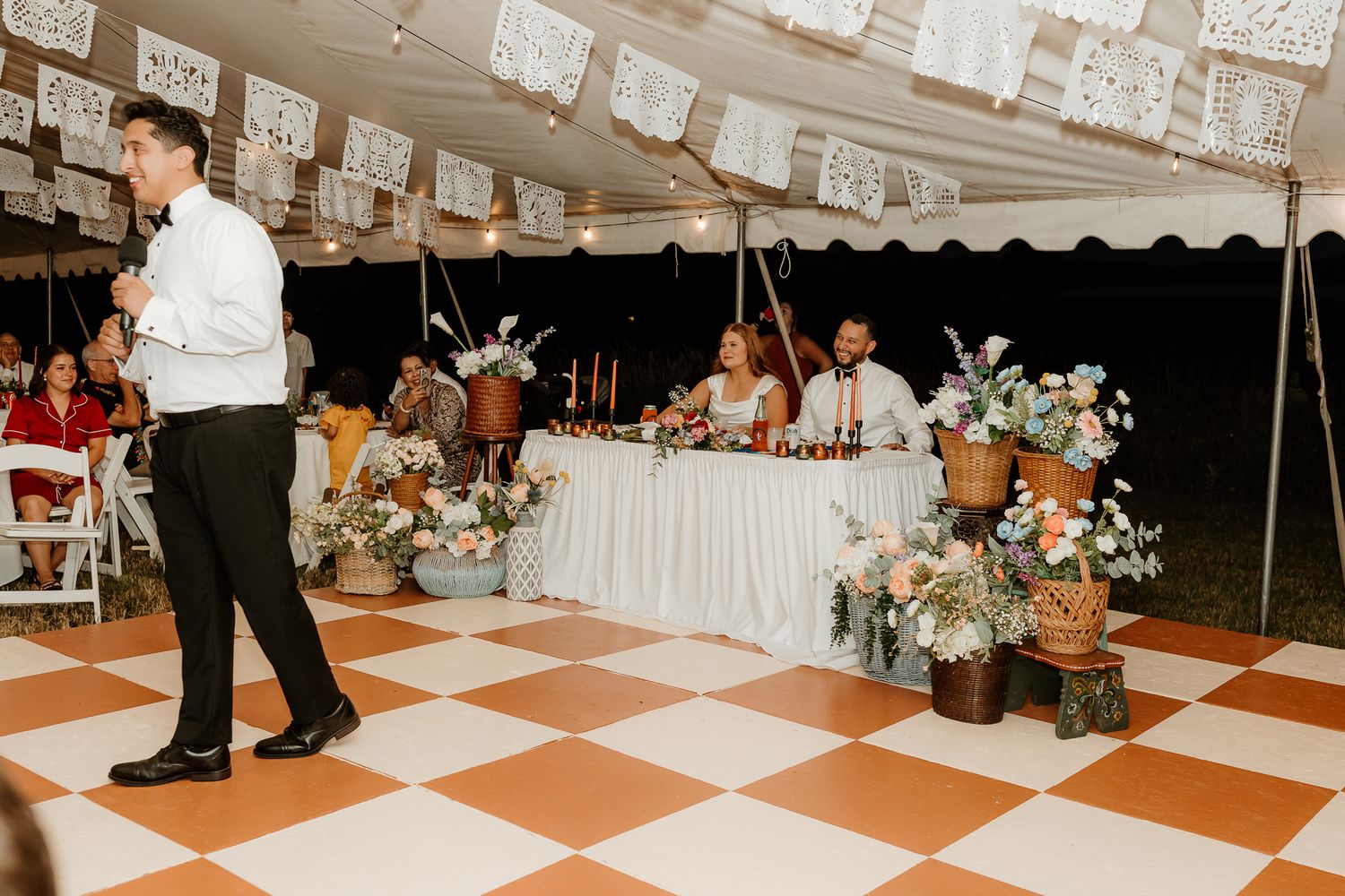 Elegant wedding reception under a tent with checkered dance floor, floral arrangements, and guests enjoying a speech.