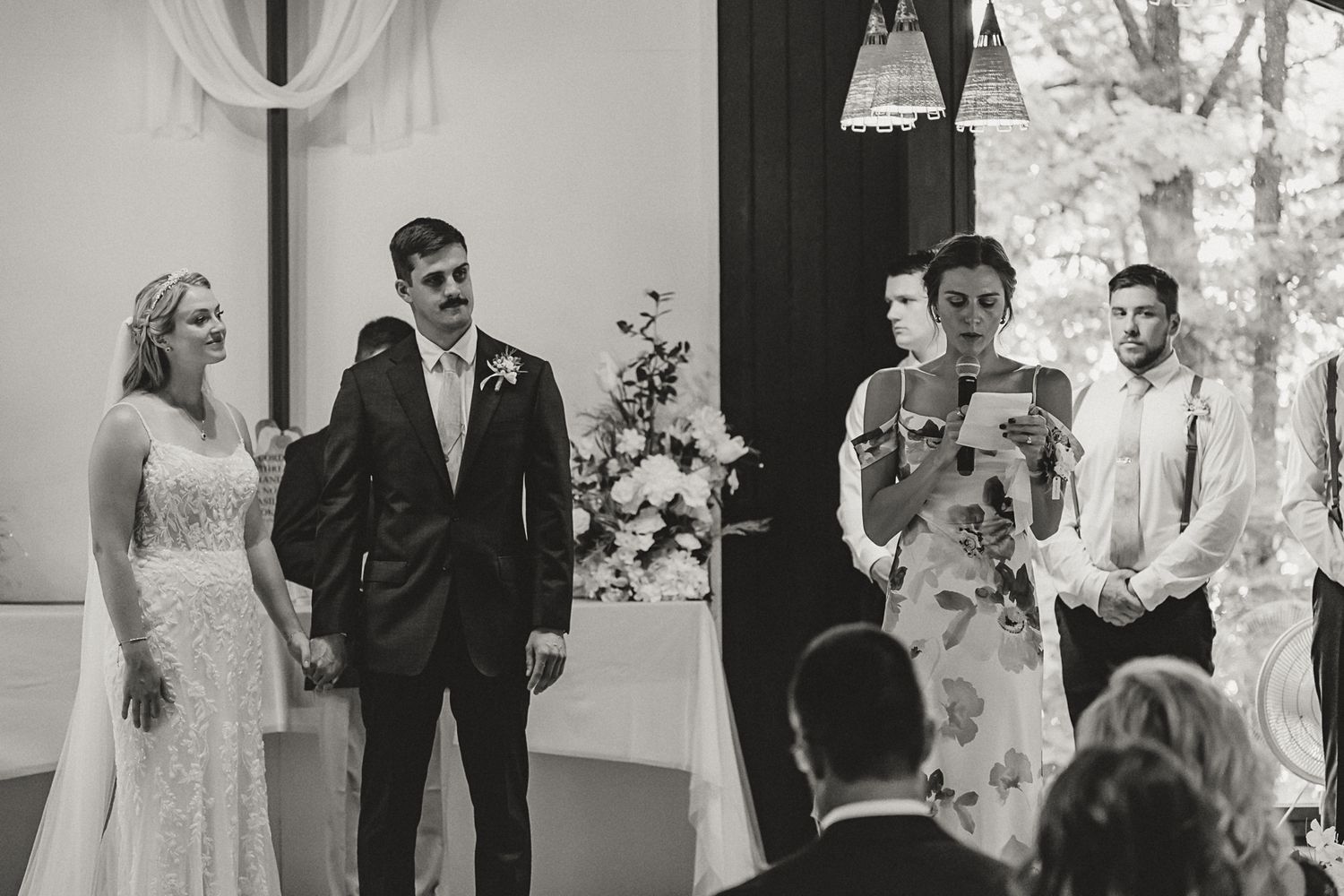 Black and white wedding ceremony photo with bride, groom, and officiant reading vows indoors.