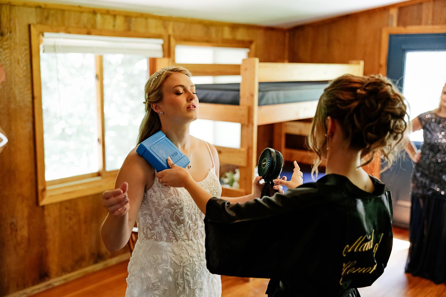 Bride in white lace wedding dress being helped by bridesmaid in rustic cabin getting-ready room.