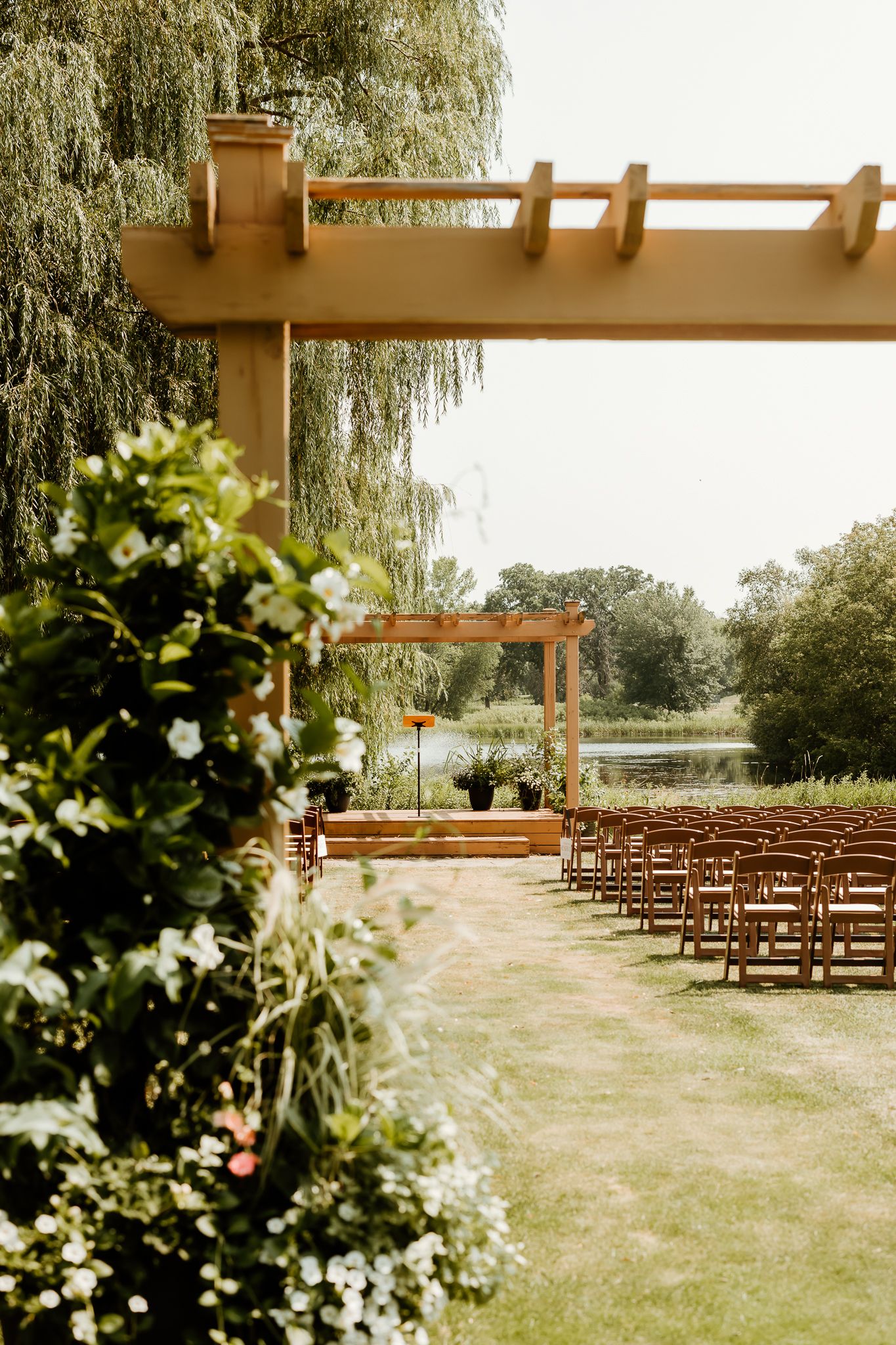 Outdoor wedding ceremony setup with wooden chairs, pergola arch, floral arrangements, and a serene pond backdrop.