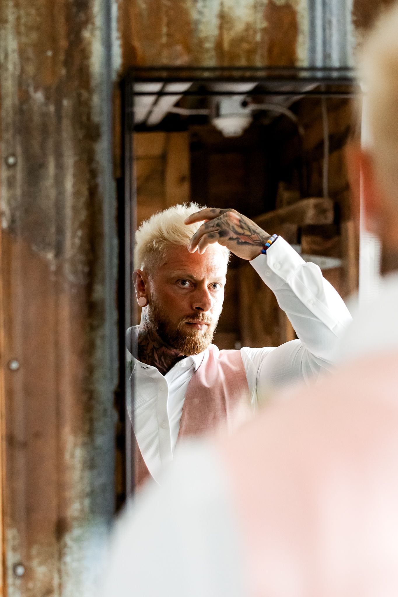 Tattooed man with blonde hair adjusting his hair in a mirror, wearing a white shirt in an industrial setting.