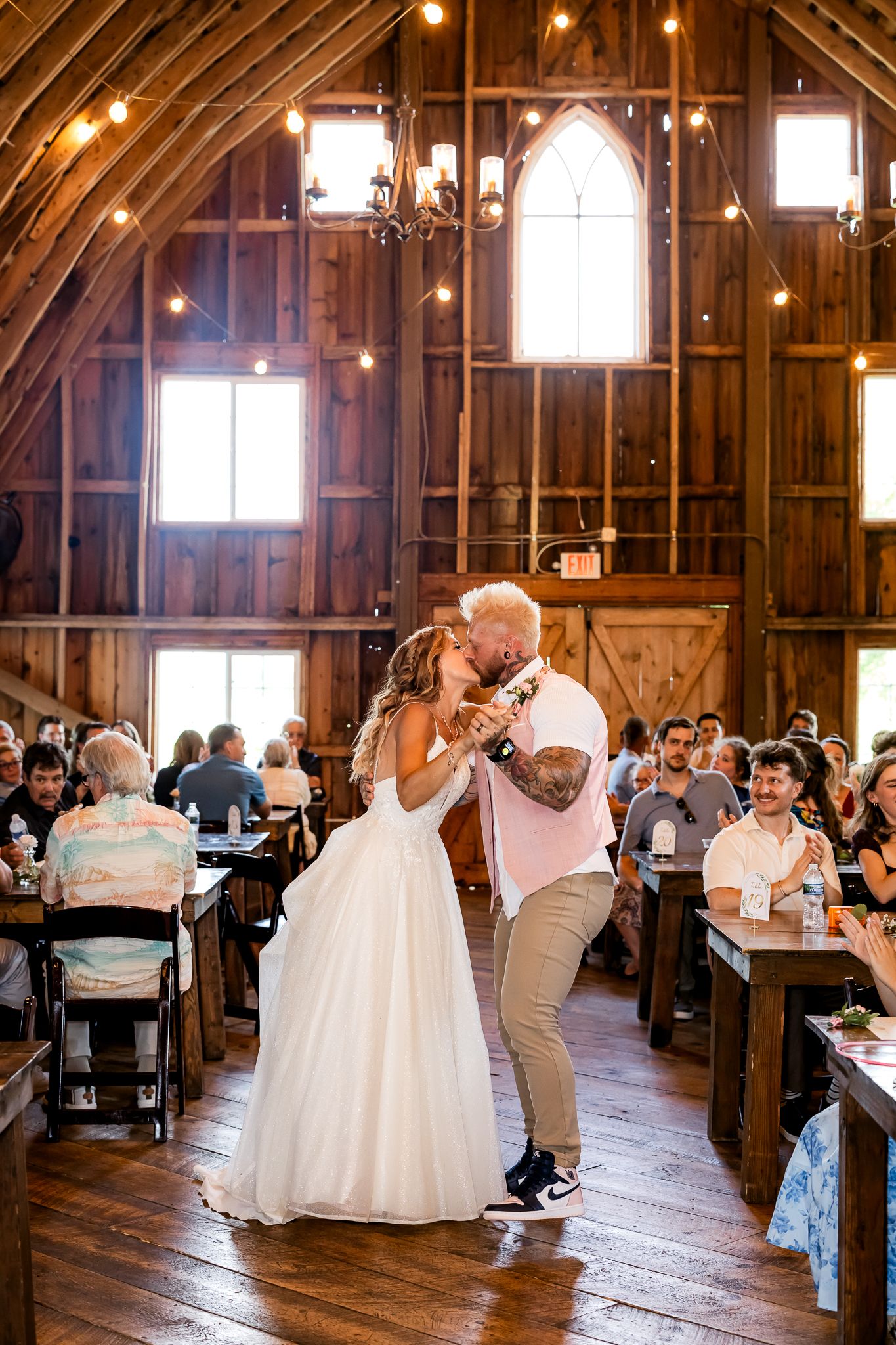 Bride and groom share first dance kiss in rustic barn wedding venue with string lights and wood walls.