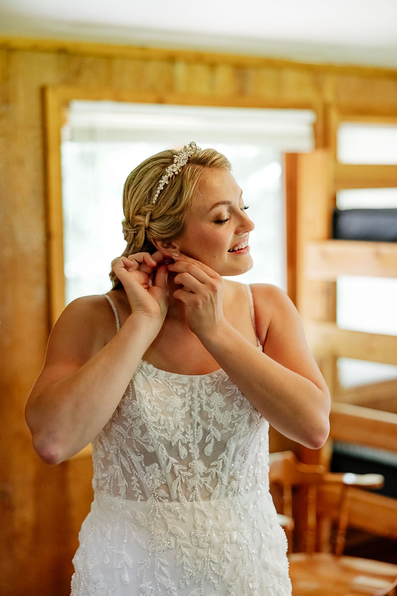 Bride in lace wedding gown adjusting earring, wearing floral headpiece, smiling in rustic wooden room.