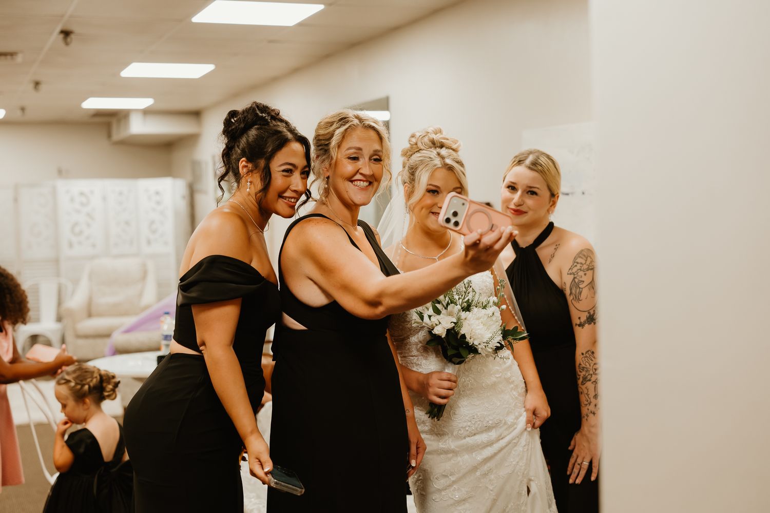 Bride in white gown takes selfie with three bridesmaids in black dresses, holding bouquet in bright room.