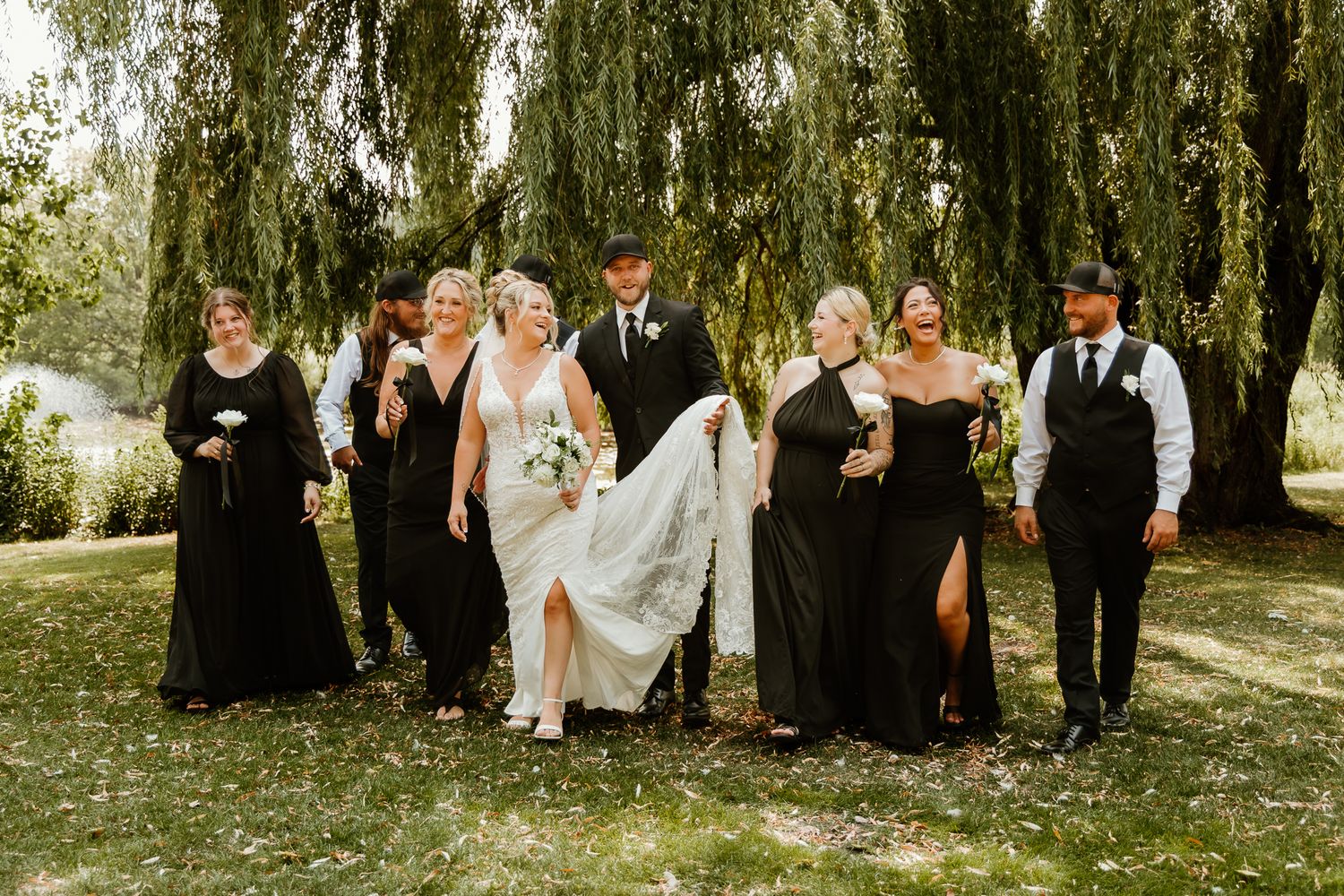 Wedding party walks outdoors under willow trees, bride in white gown with bridal party in elegant black attire.
