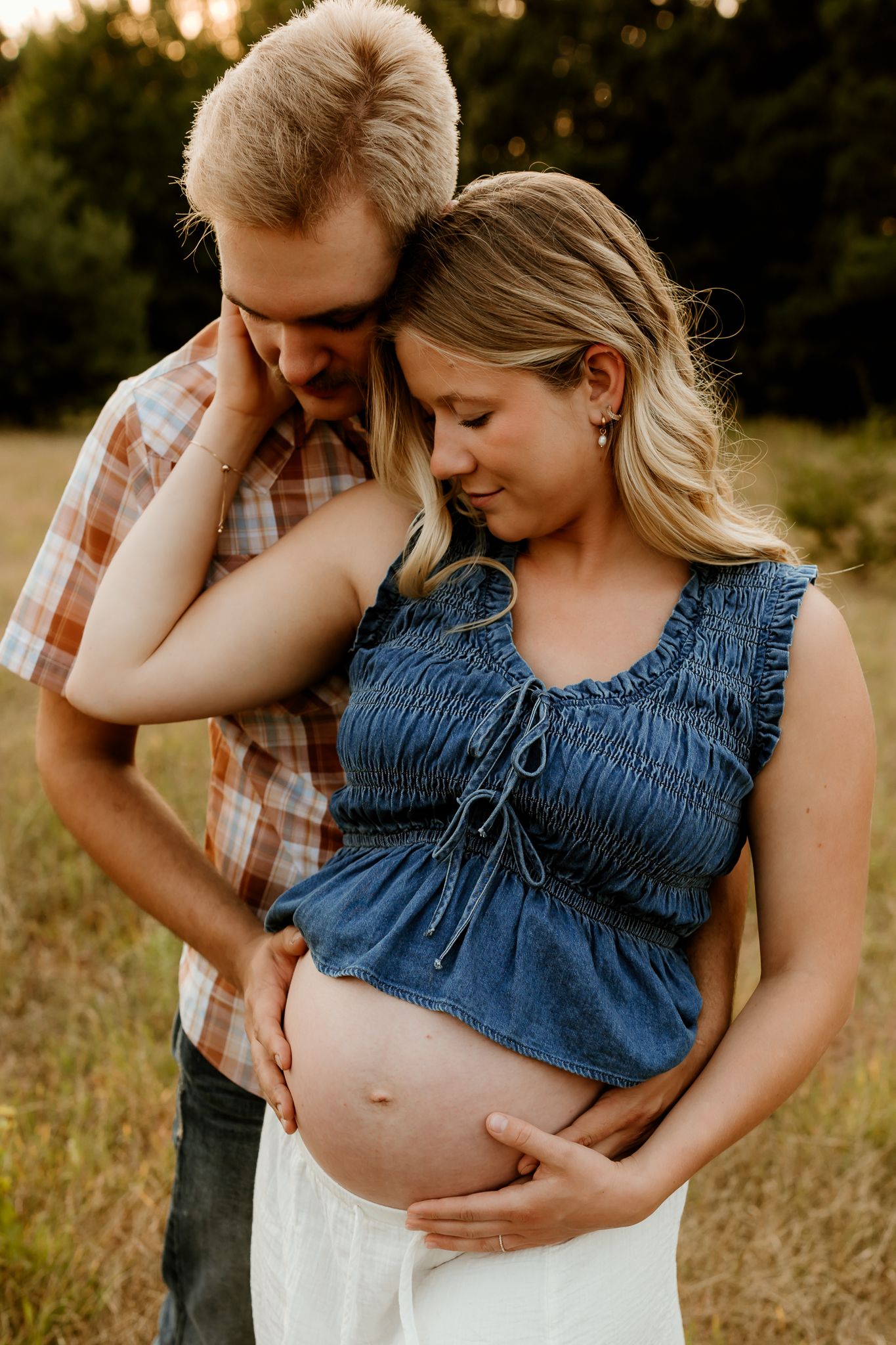 Couple sharing a tender moment outdoors, woman in denim top showing baby bump, man in plaid shirt embracing her.