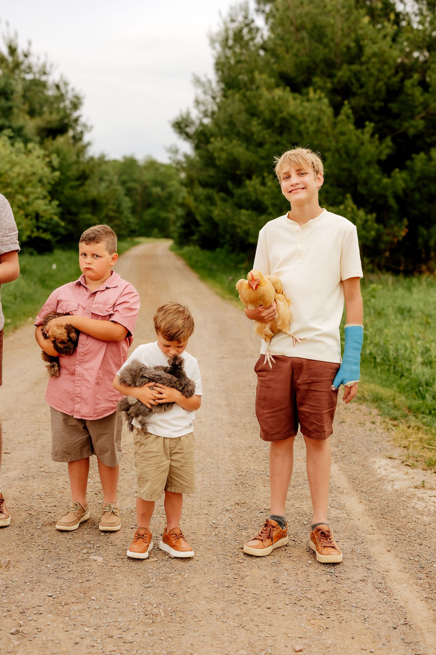 Three boys smile on a rural dirt path holding chickens and small animals in coordinating casual outfits.