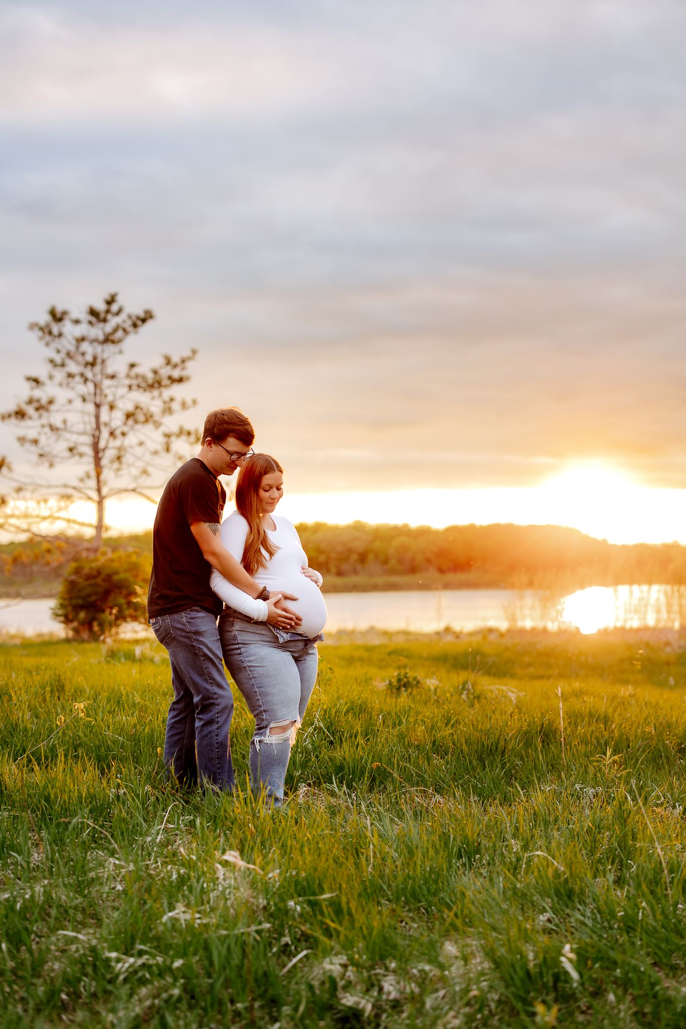 Couple cradling a baby bump at golden sunset by a lake, standing in a lush green field.