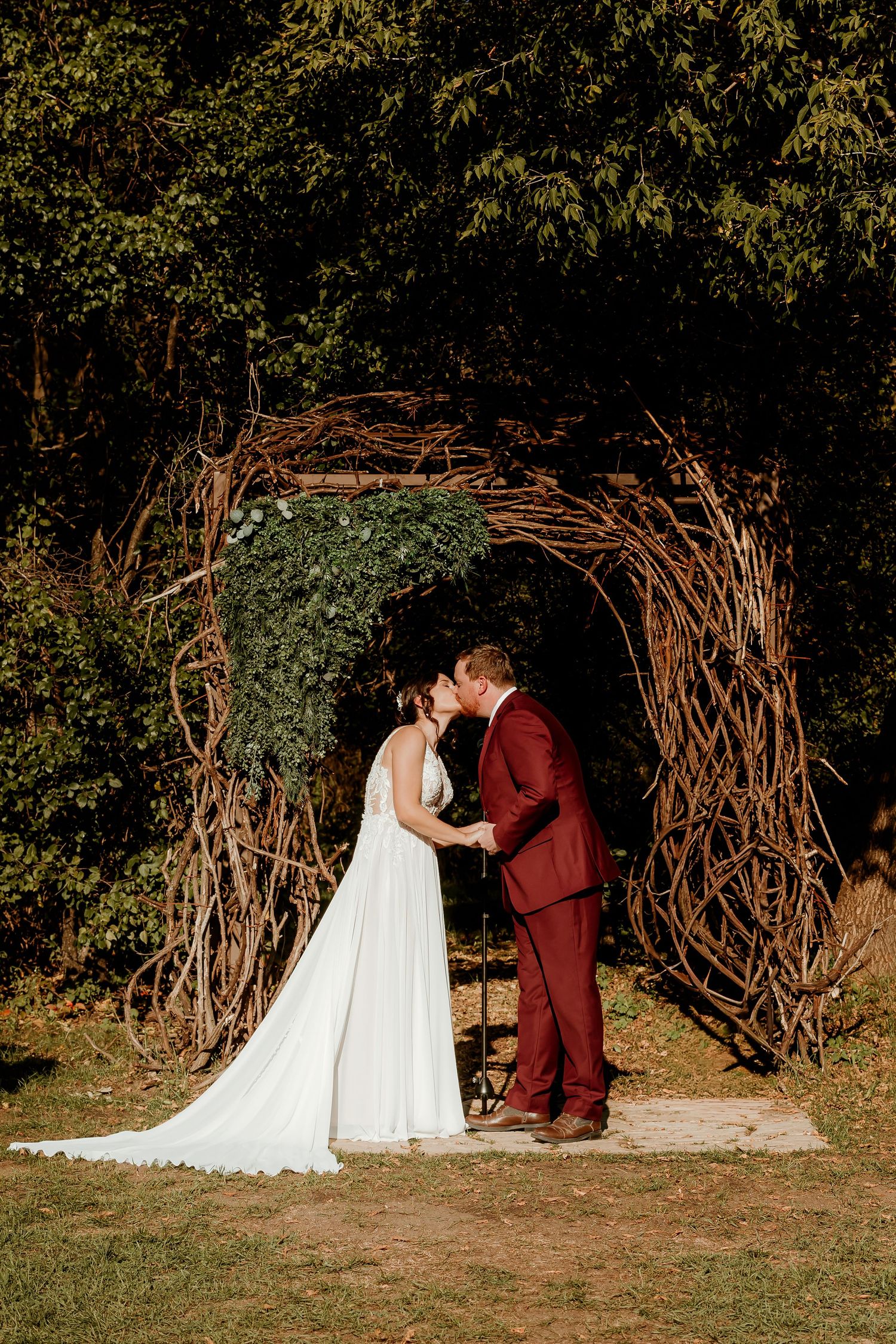Bride in a white gown and groom in a burgundy suit kiss under a rustic twig wedding arch with ivy at an outdoor ceremony.