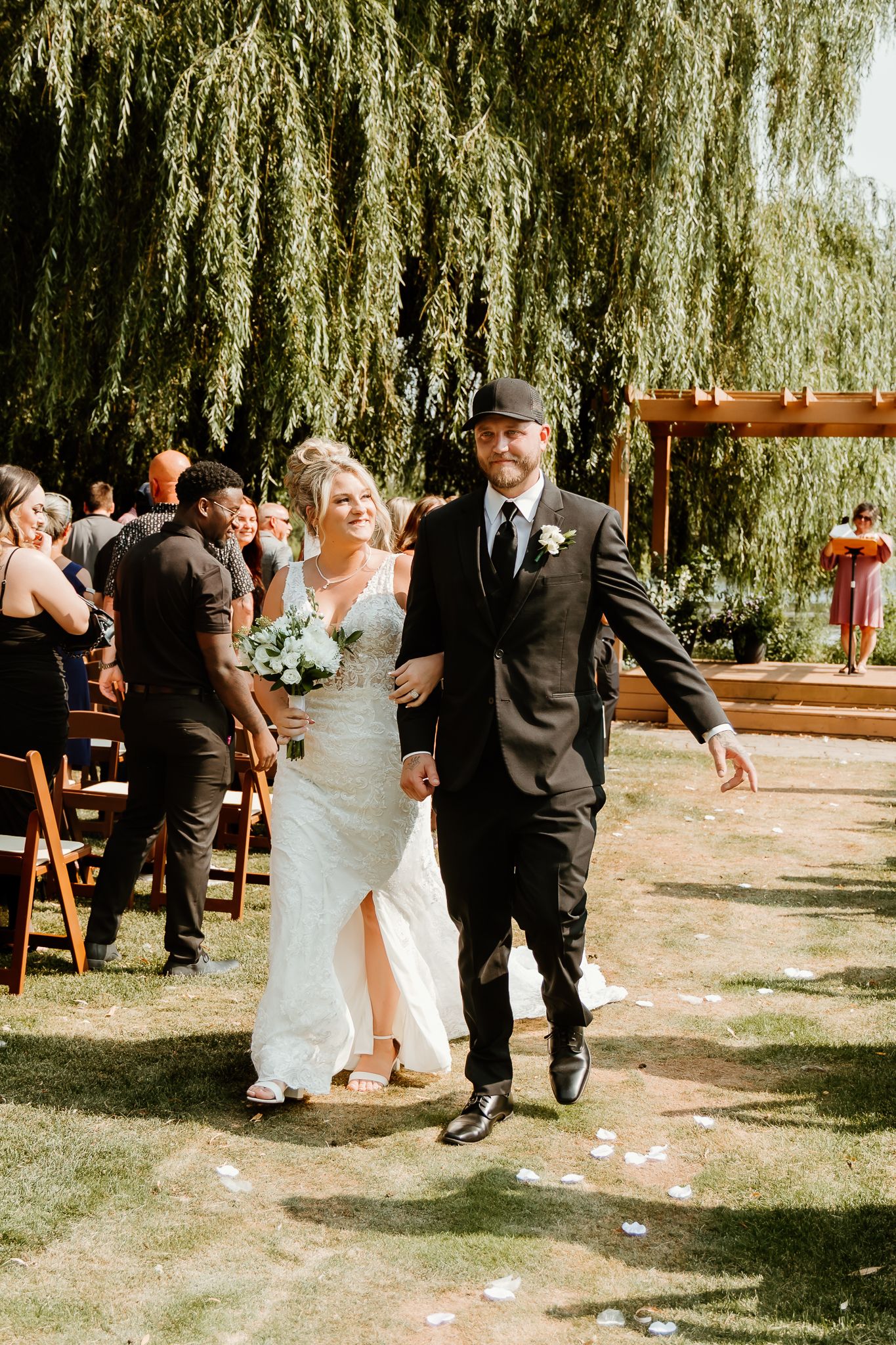 Bride and groom walk down outdoor aisle under weeping willow tree after wedding ceremony on sunny day.